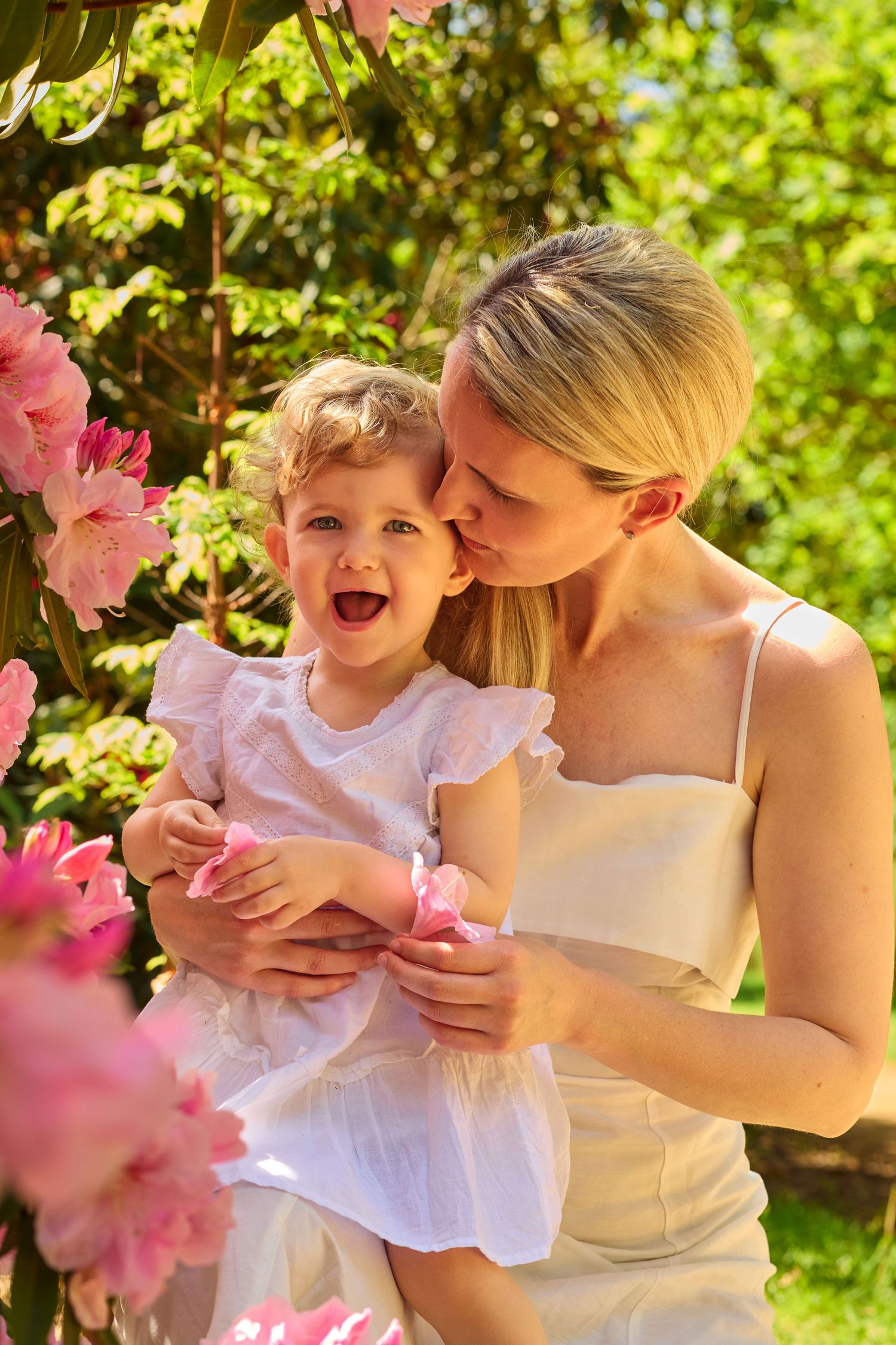 Mother kissing her laughing toddler daughter among pink blooming flowers in a sunny garden.