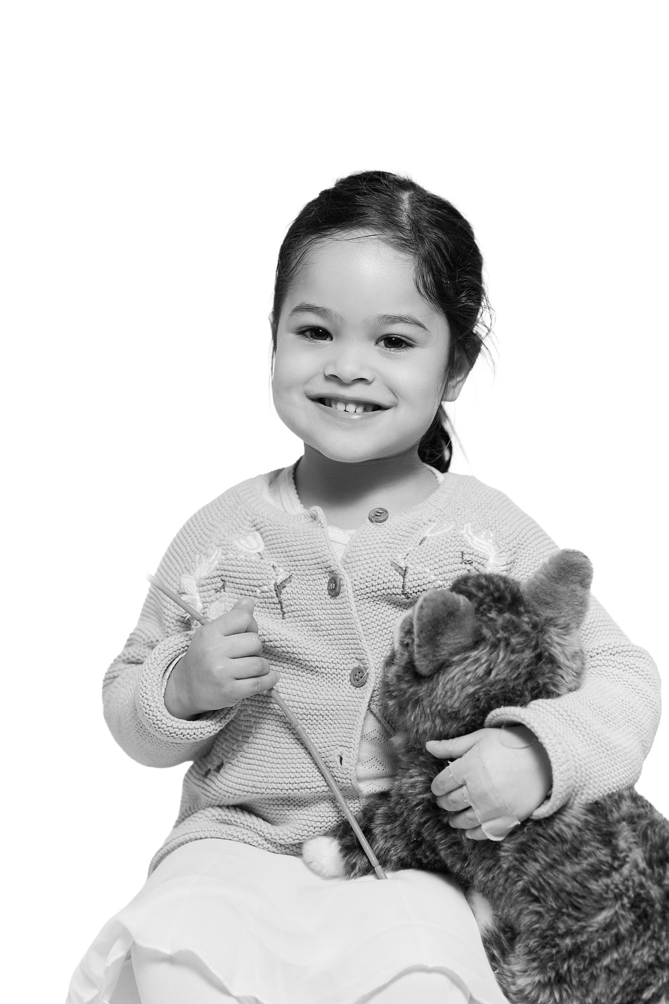 Happy toddler girl in knit cardigan hugging stuffed animal toy, black and white studio portrait.
