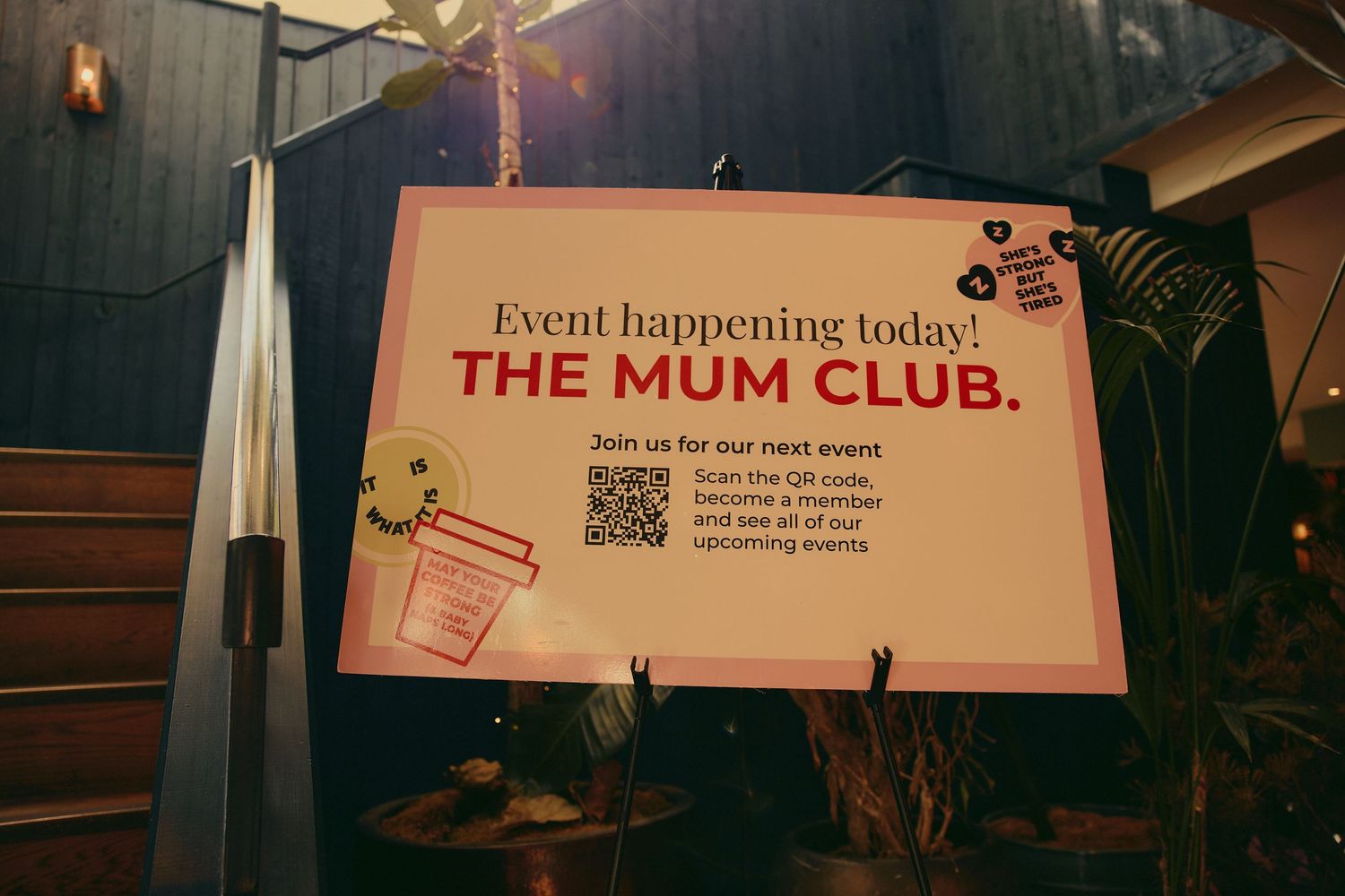 A sign for The Mum Club event with QR code displayed outdoors at a venue.