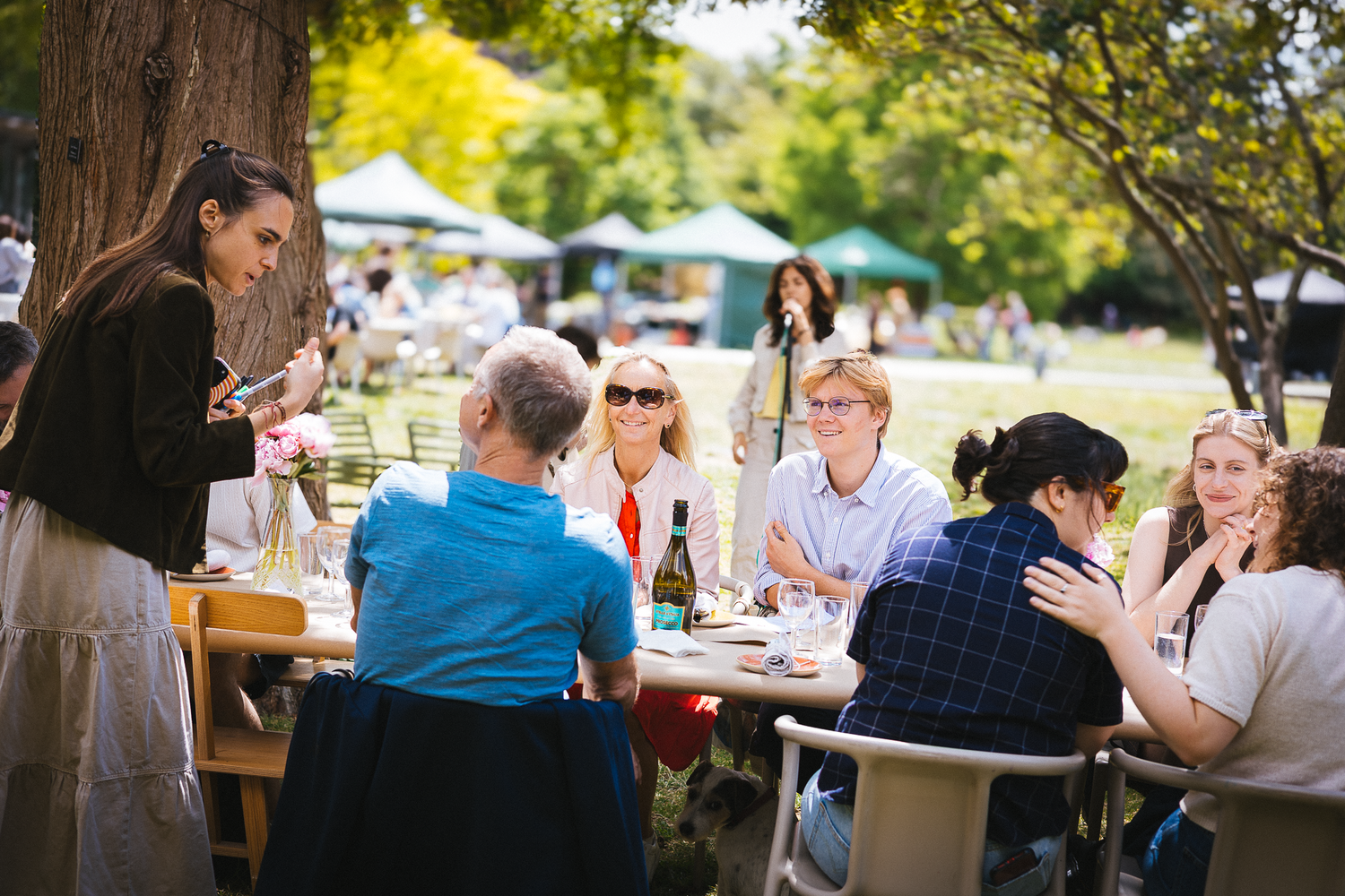 Guests mingle at outdoor tables under trees during a daytime event.