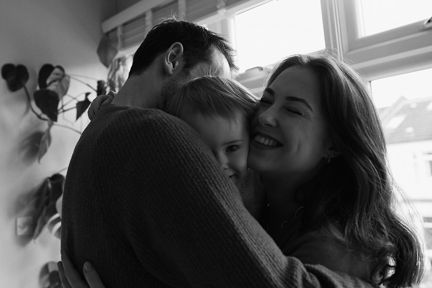 Black and white photo of a happy family sharing a warm embrace, mother smiling as parents hug young child.