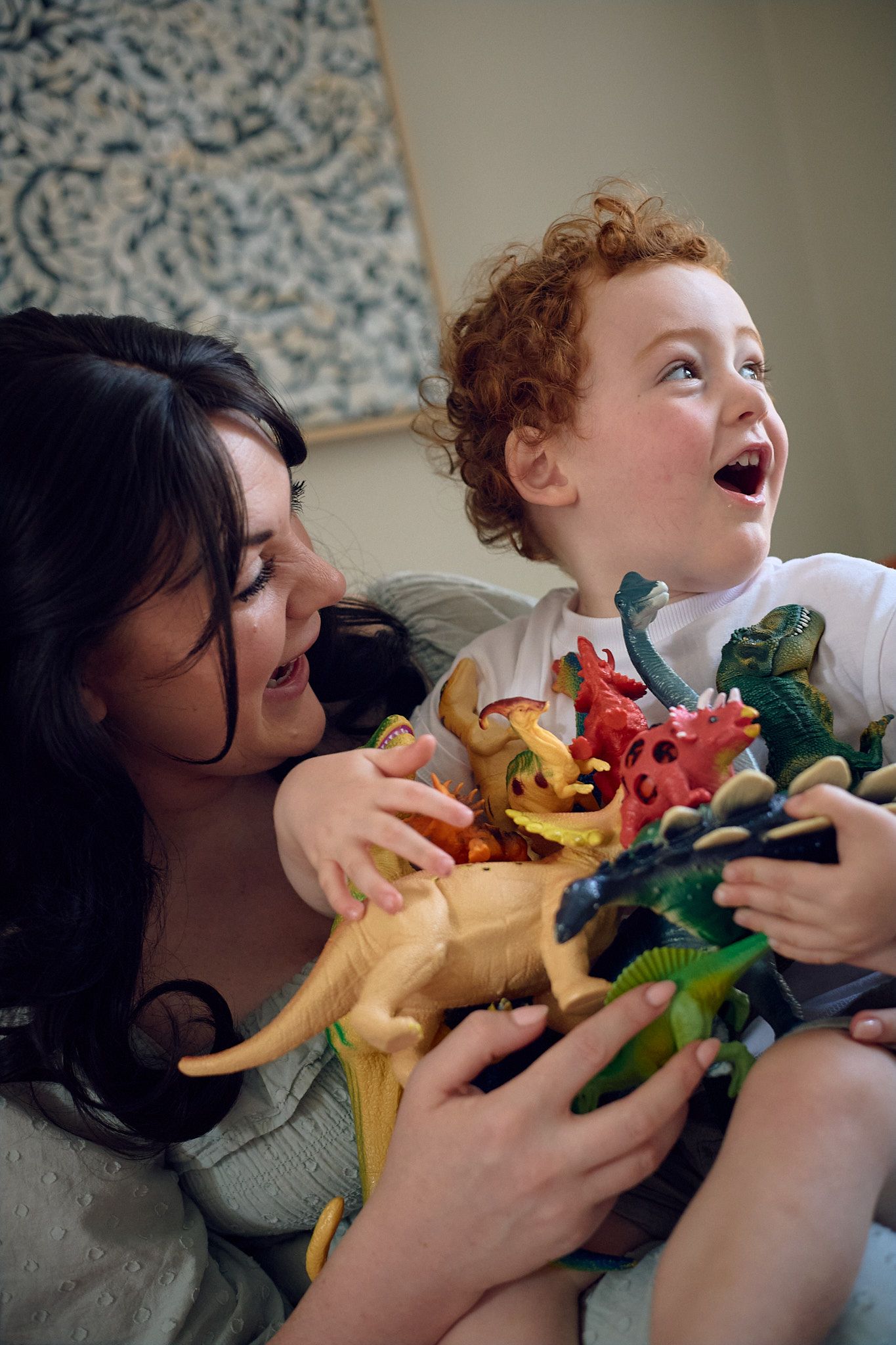 A joyful moment playing with colorful dinosaur toys on a couch with a patterned wall in the background.