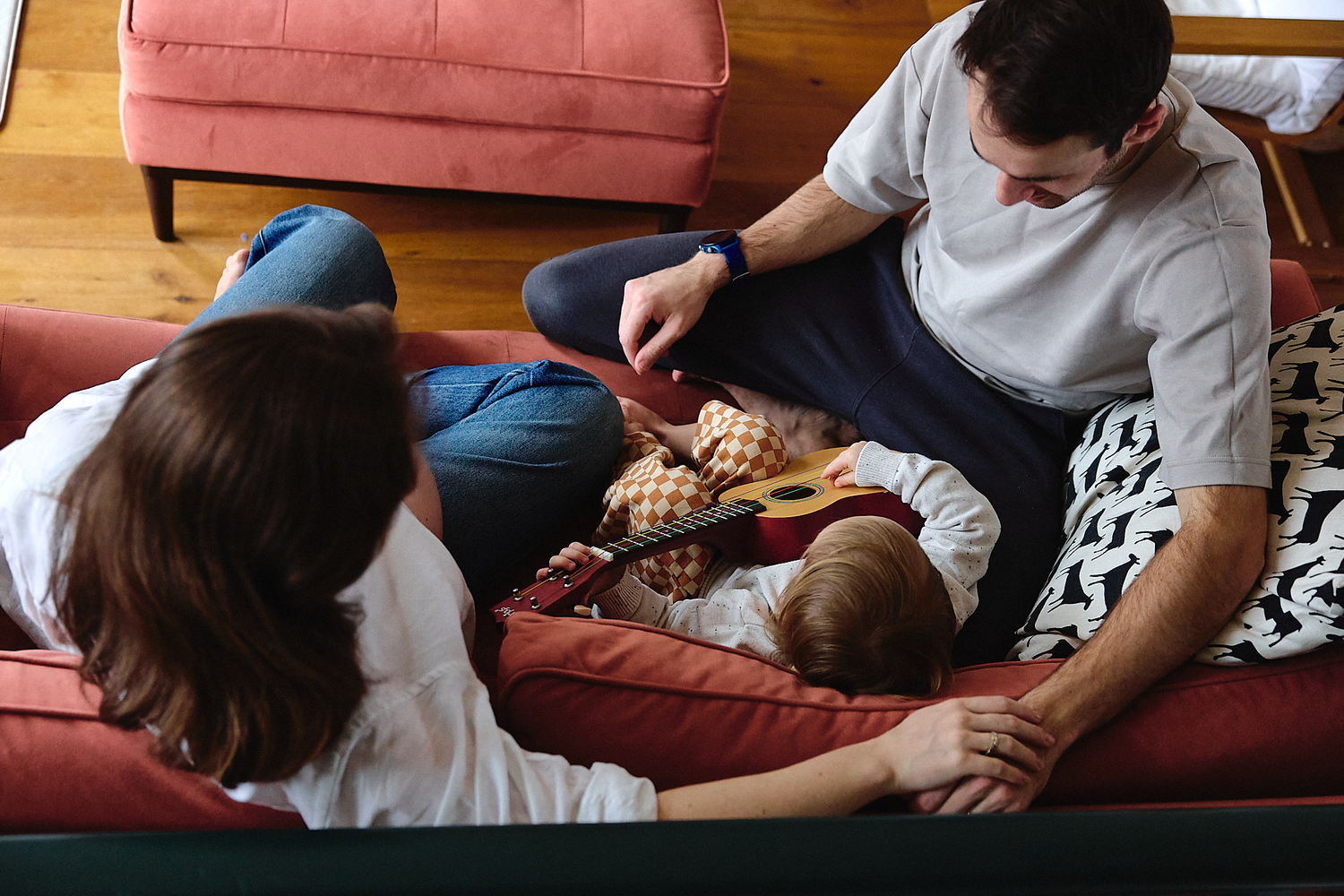 Overhead view of family relaxing on red sofa with young child between parents, sharing snacks.