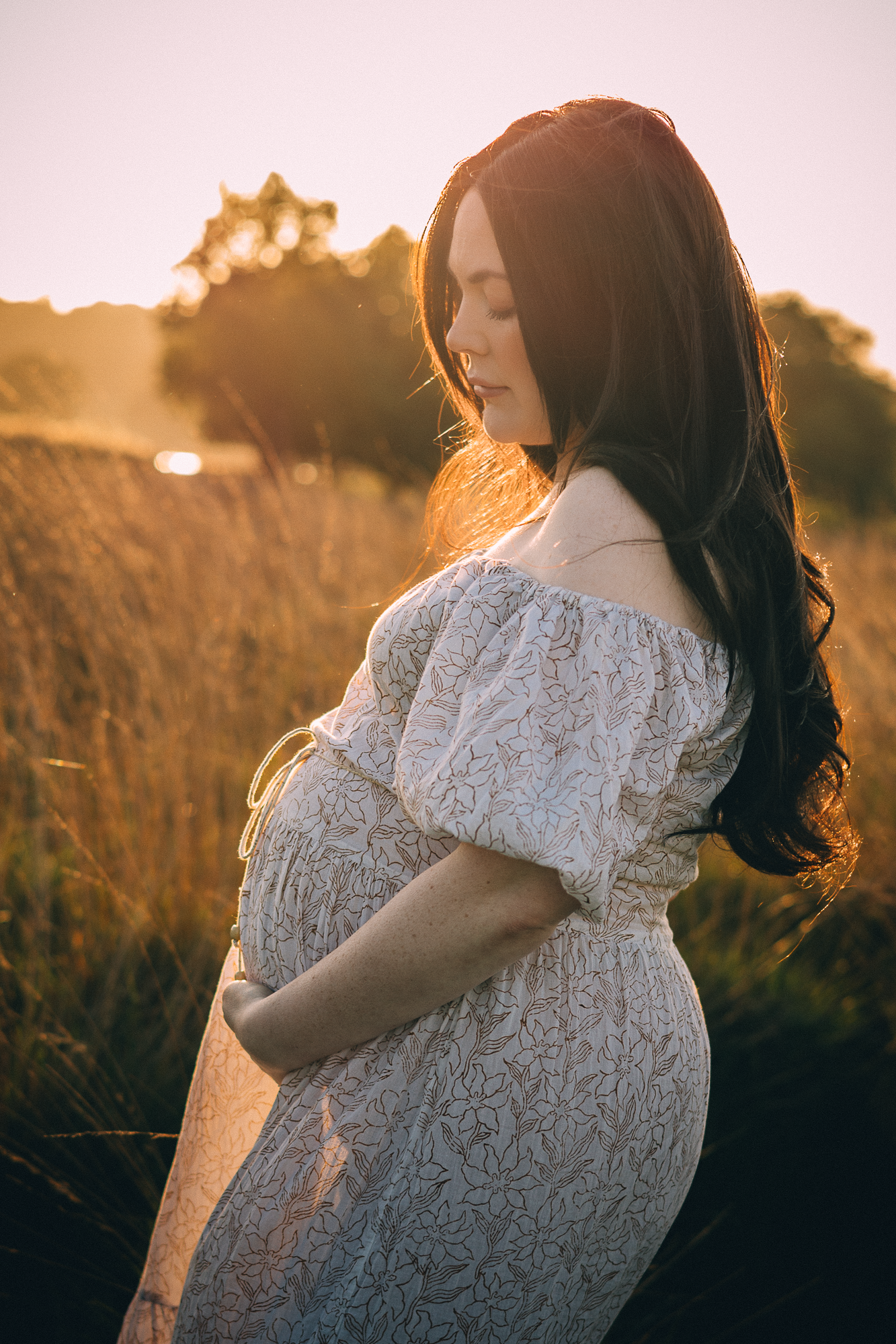 Dreamy backlit maternity photo in a field at golden hour showing lace dress details.