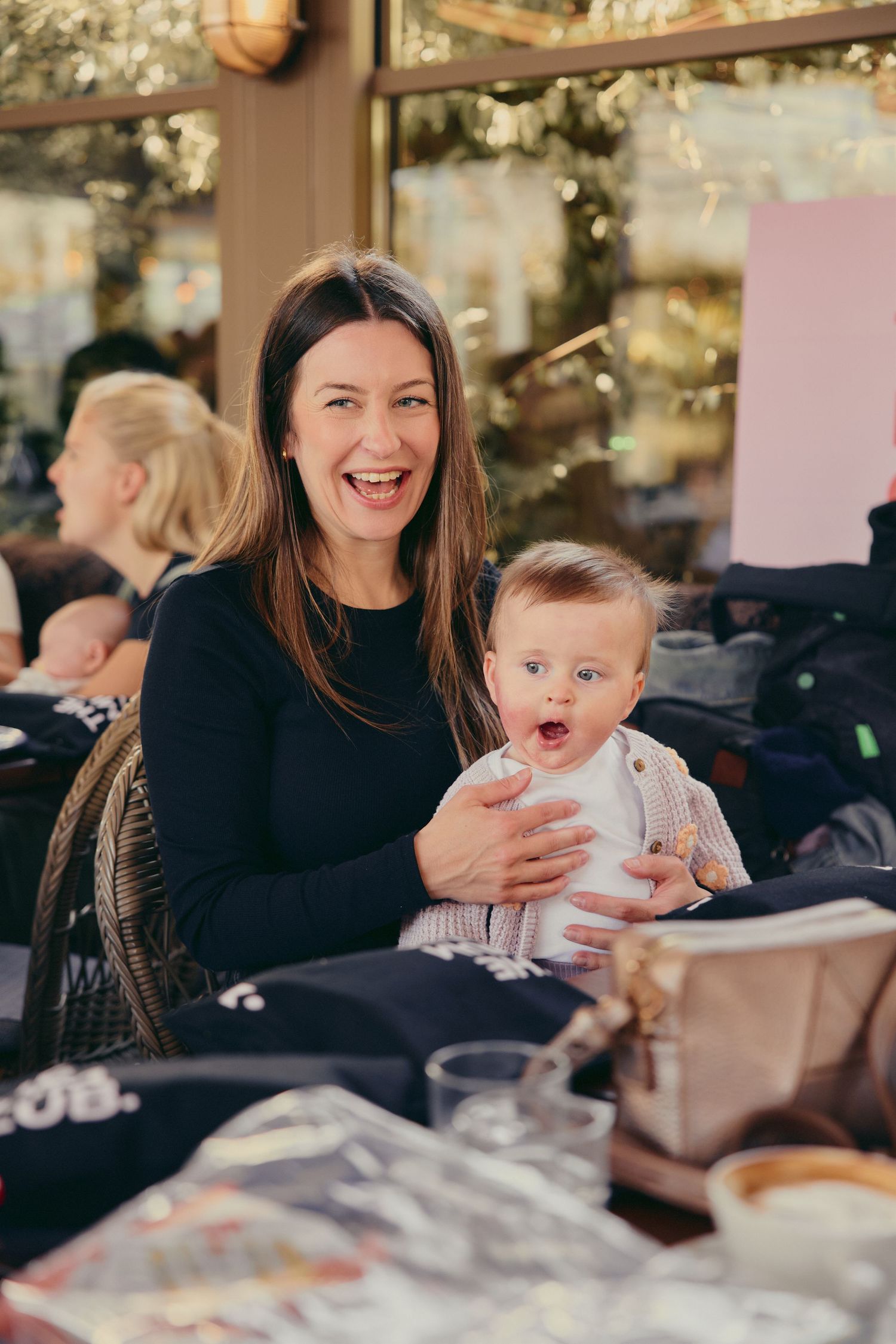 A happy brunette mother laughing while holding her wide-eyed baby at a restaurant gathering.