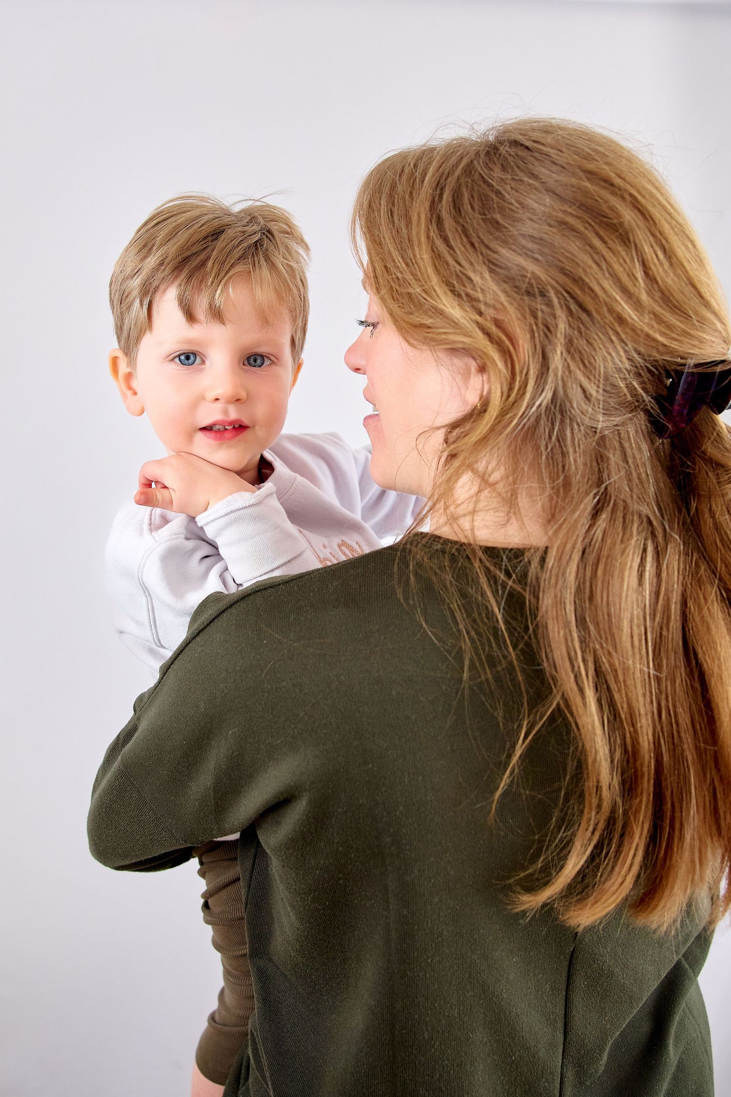 A woman with long blonde hair holds a young blue-eyed boy in a white shirt, smiling at each other tenderly.