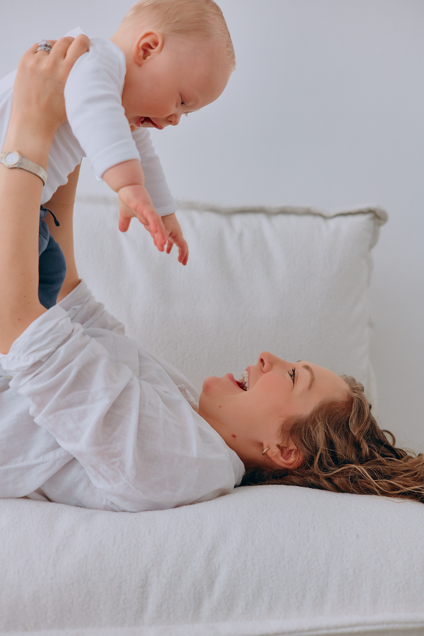 A joyful mother lying on a white bed, lifting her smiling baby in the air during playful bonding time.