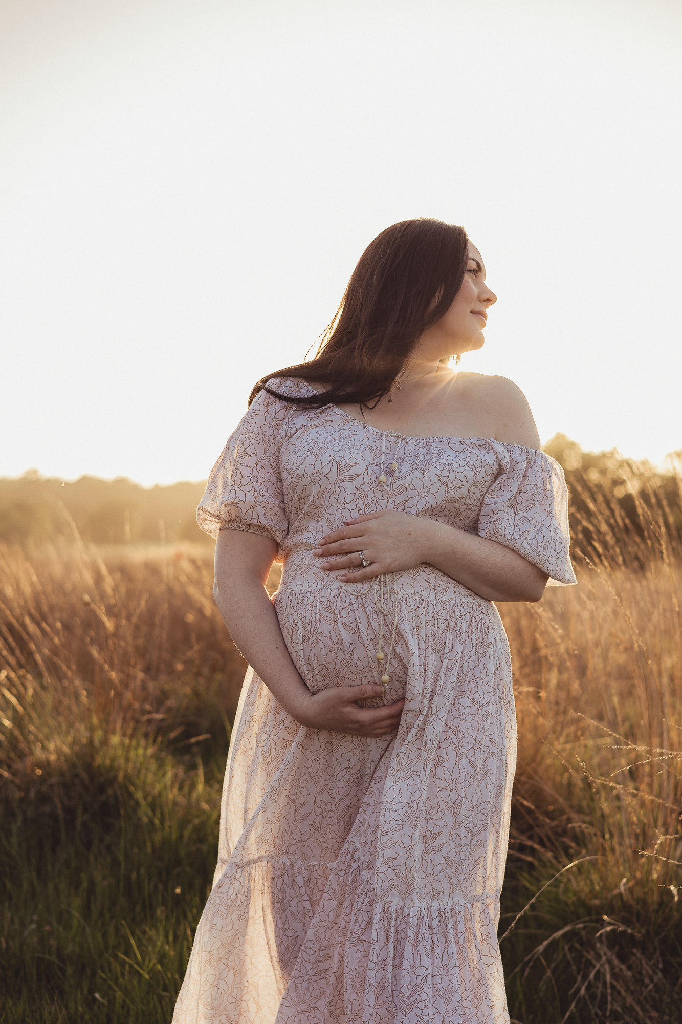 Maternity portrait of woman in lace dress standing in sunlit field at golden hour.