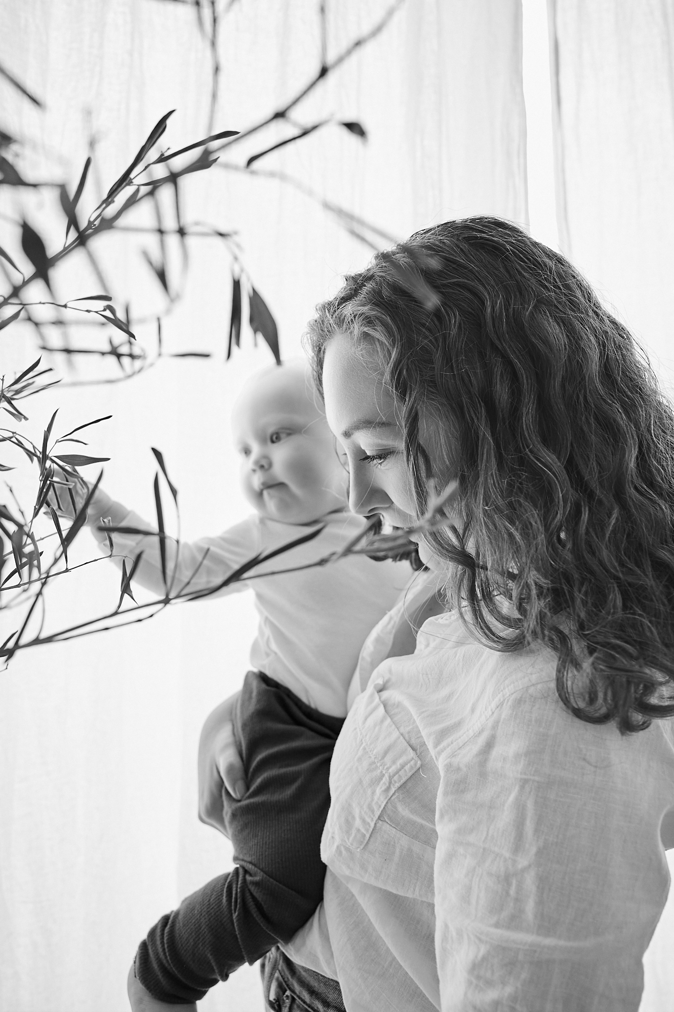 Black and white photo of a curly-haired mother tenderly holding her baby near leafy branches by a bright window.
