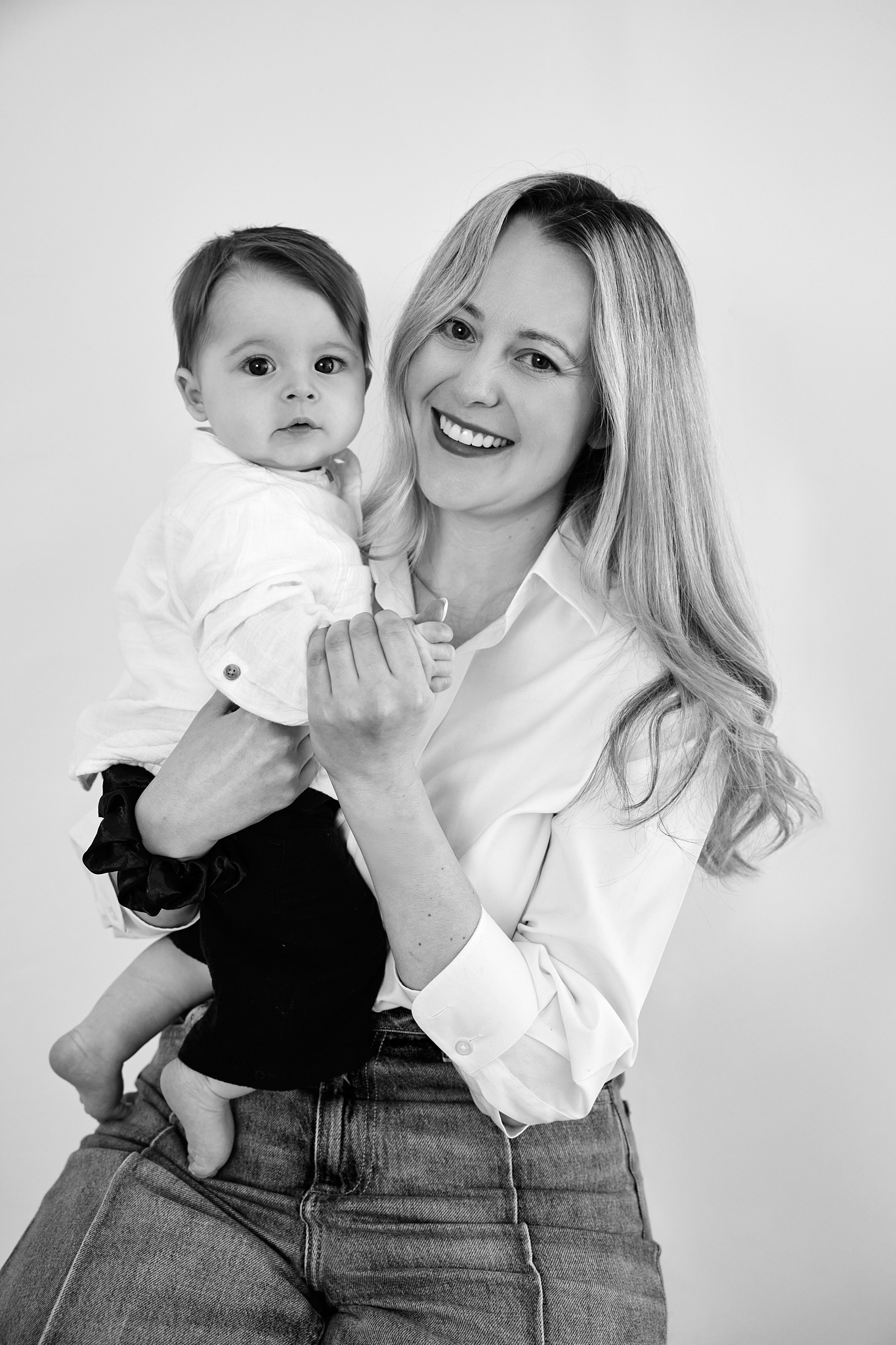 Black and white photo of a smiling blonde woman holding a cute baby dressed in white shirt and dark pants.