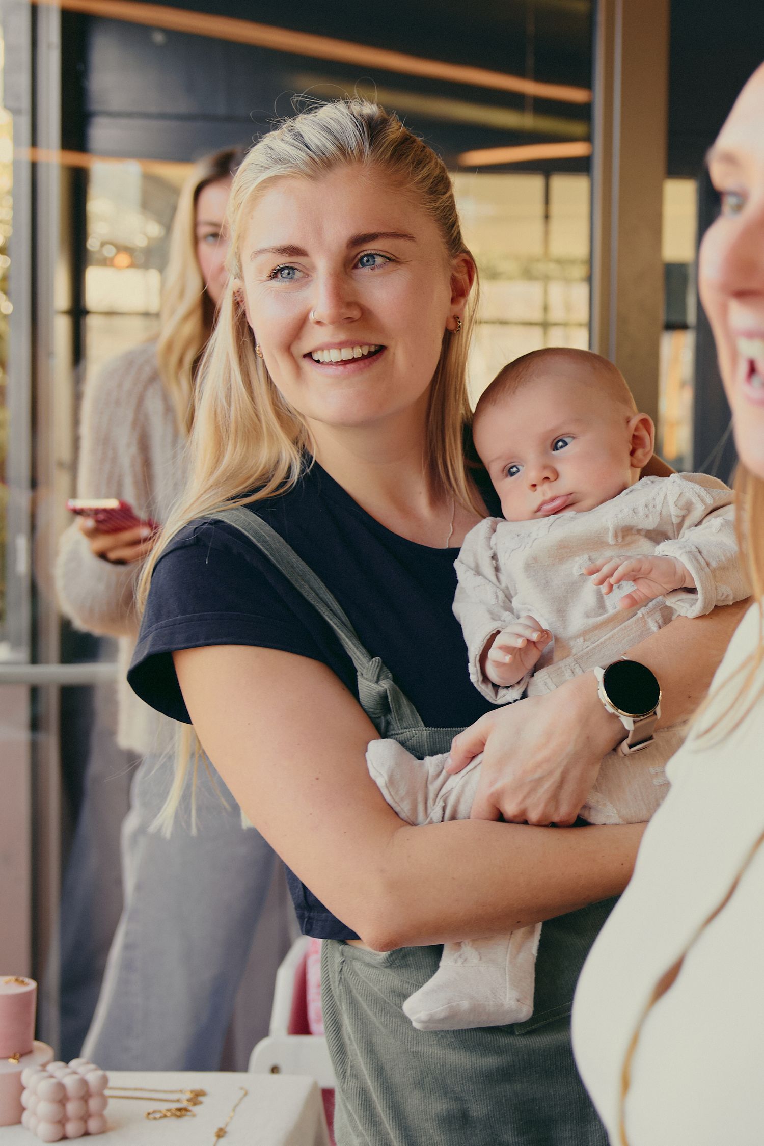 Smiling blonde woman holding a newborn baby at a social gathering indoors.
