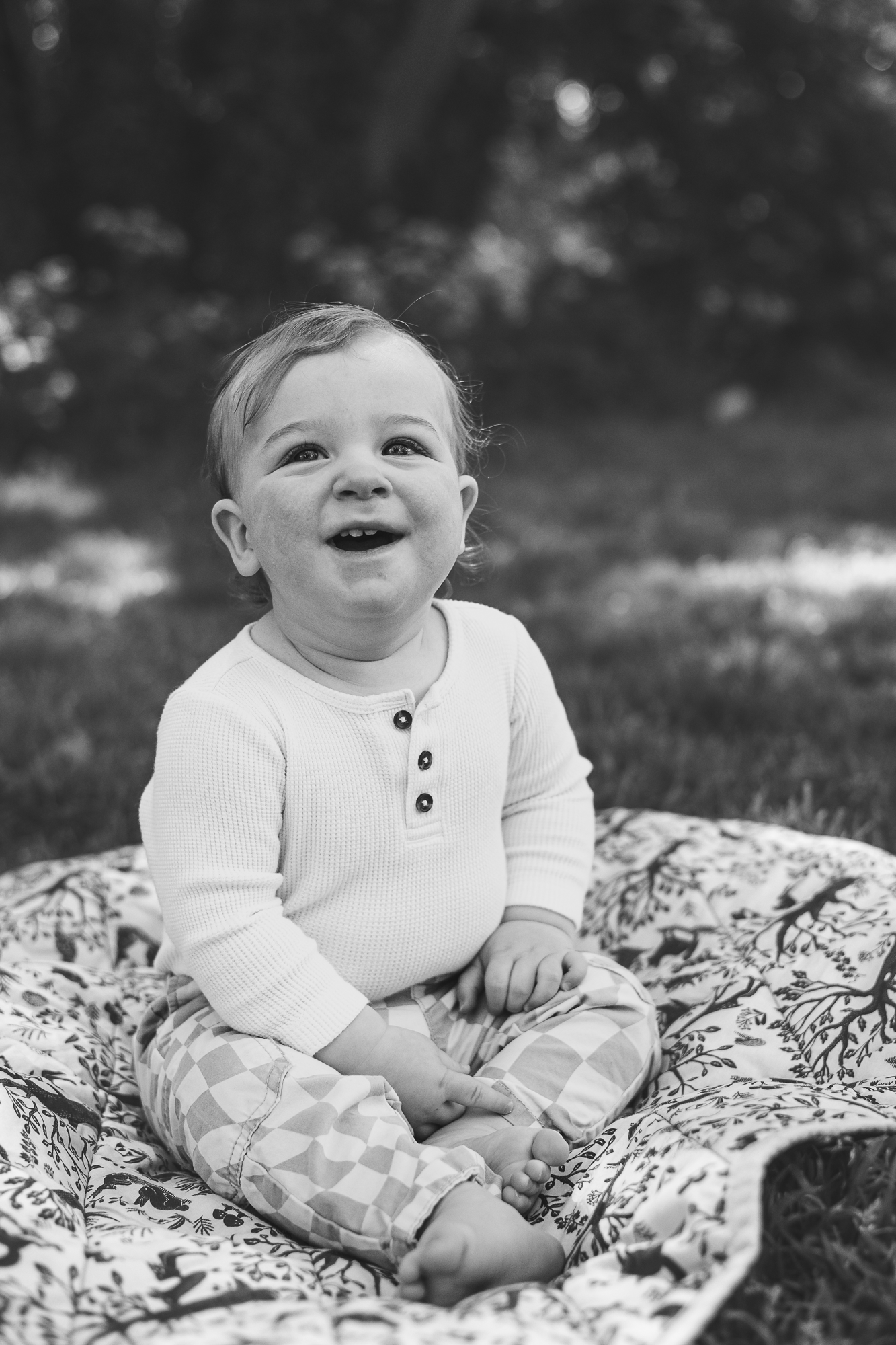 Baby in white henley top sits on patterned blanket outdoors with joyful expression.