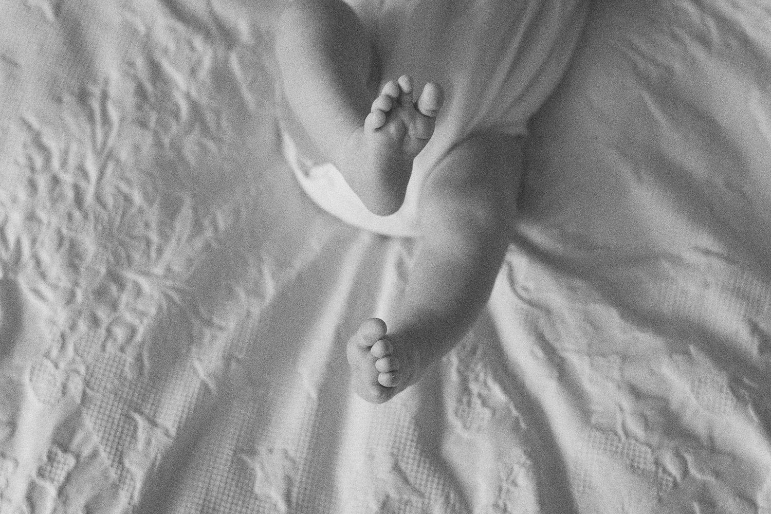 Black and white photo of newborn baby feet and legs resting on a soft white blanket.