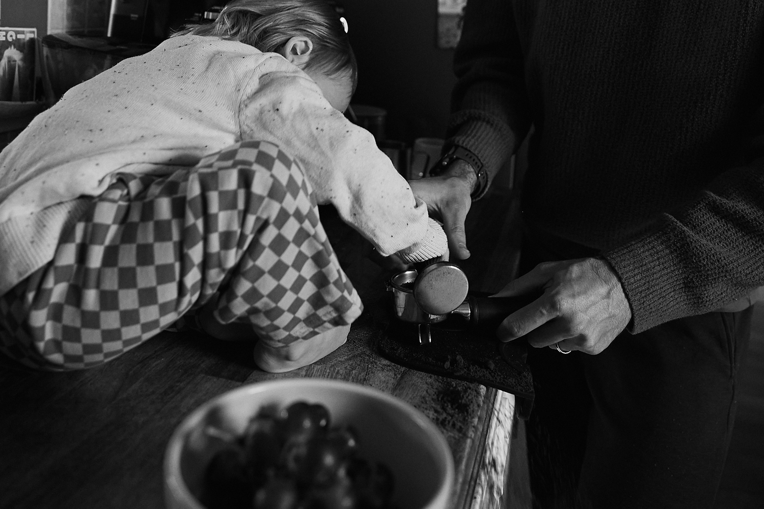 Black and white photo of child leaning over counter while adult cuts food on cutting board nearby.