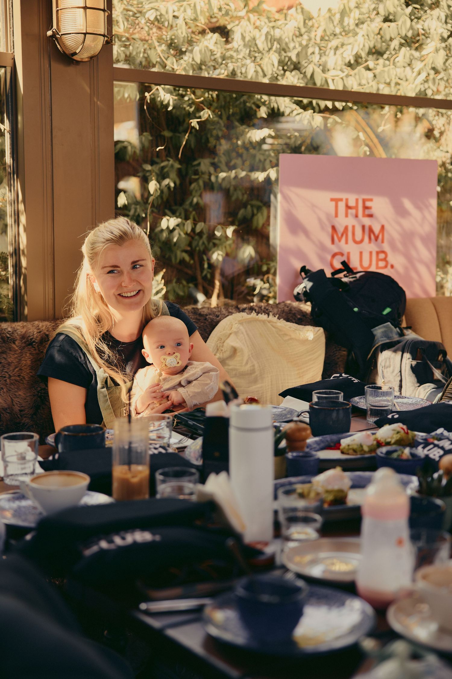 Mother holds baby at a table during The Mum Club event in a sunlit venue.