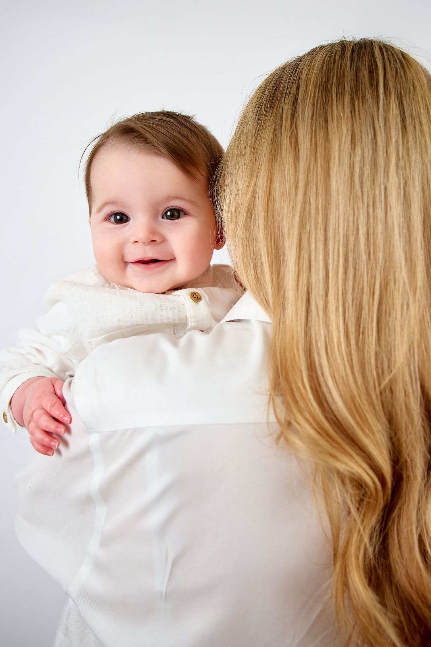 A smiling baby in white outfit being held over the shoulder of a blonde-haired mother against a white background.