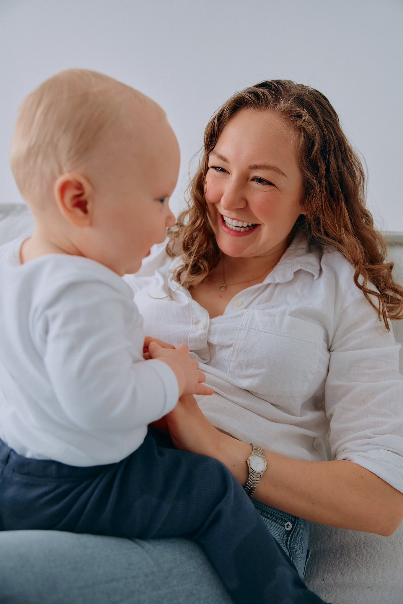 A laughing mother in a white shirt holds her baby in a bright, airy room, sharing a joyful moment together.