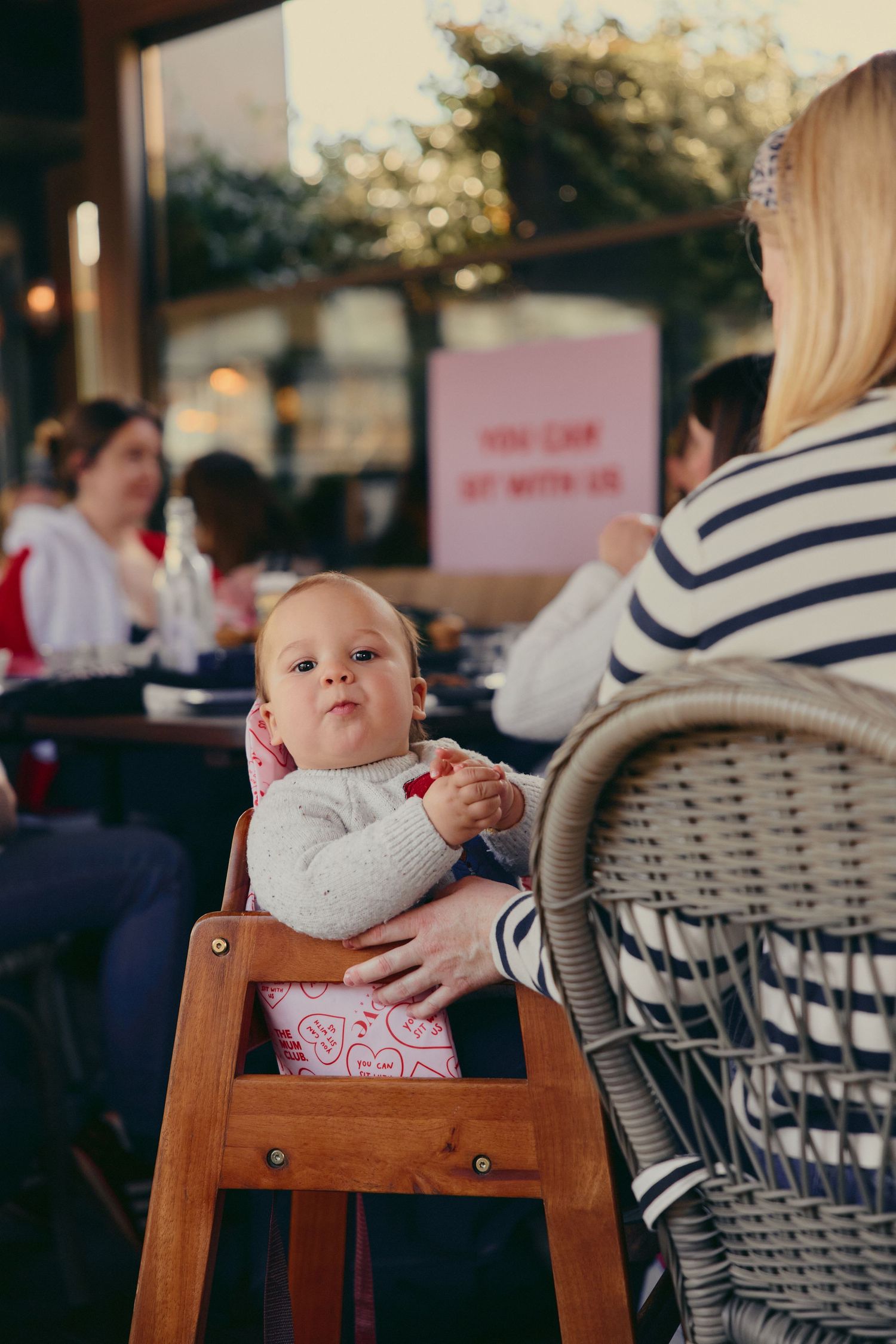 Baby in high chair at outdoor cafe with mother in striped shirt nearby.