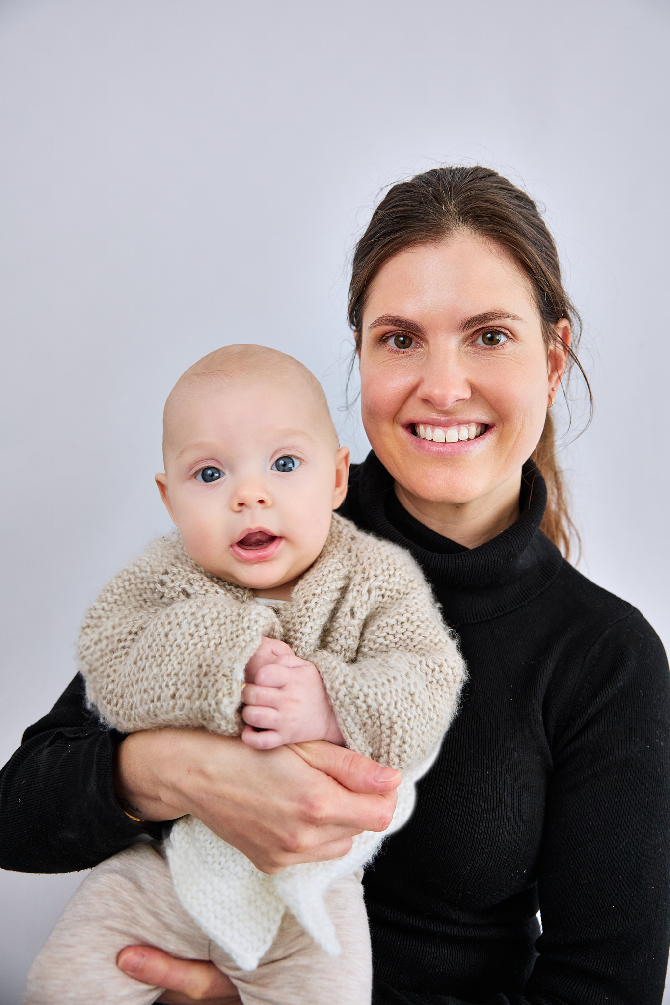 Smiling woman in black turtleneck holding a blue-eyed baby in a knit sweater against a white background.