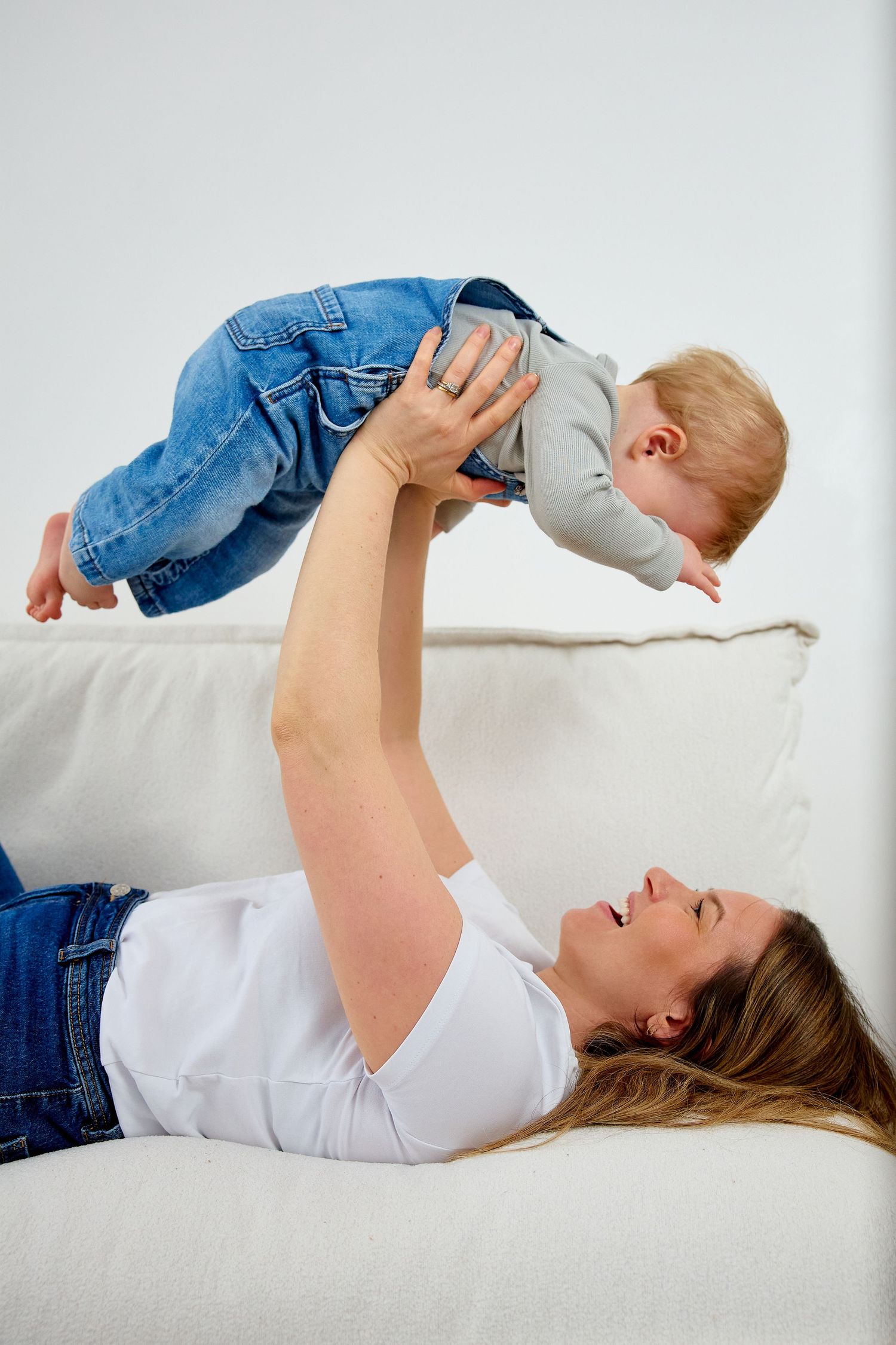 Mother lying on white sofa lifting smiling baby in jeans overhead in a playful bonding moment at home.