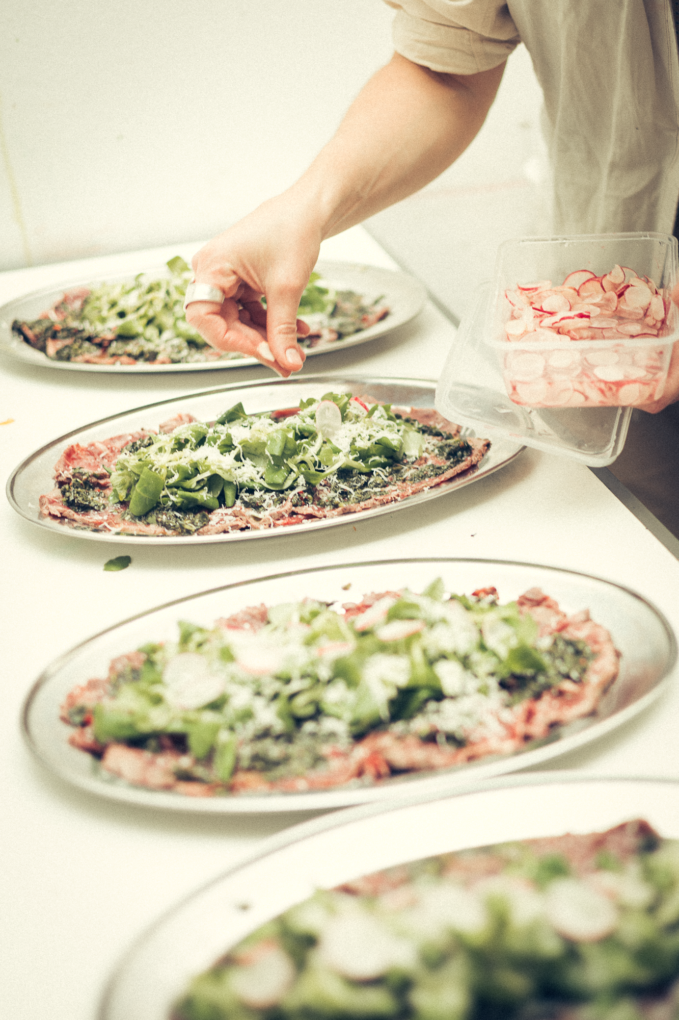 Multiple oval platters of fresh salads being garnished with greens and pink ingredients.