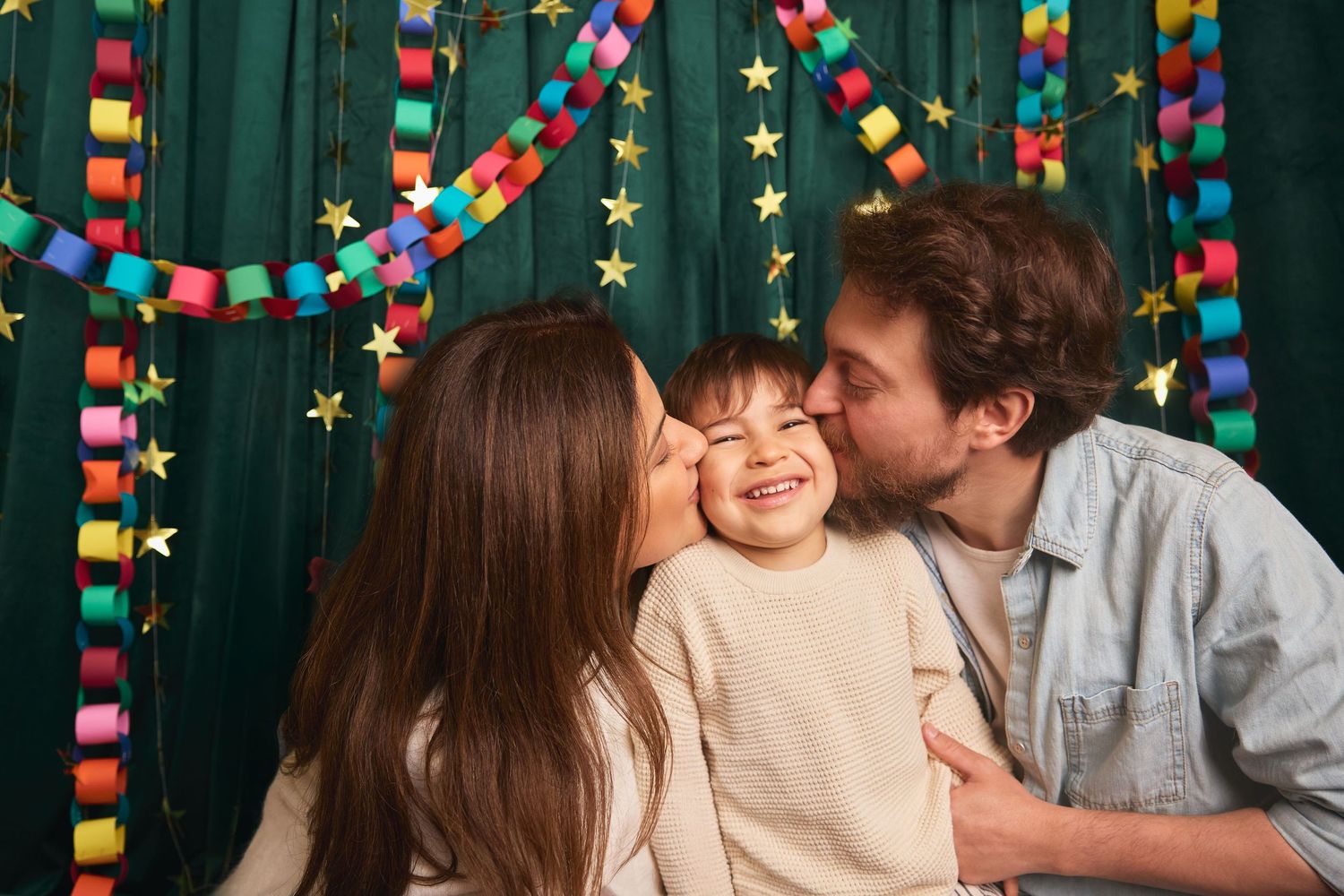 Happy family with parents kissing their smiling toddler child against colorful party decoration background.