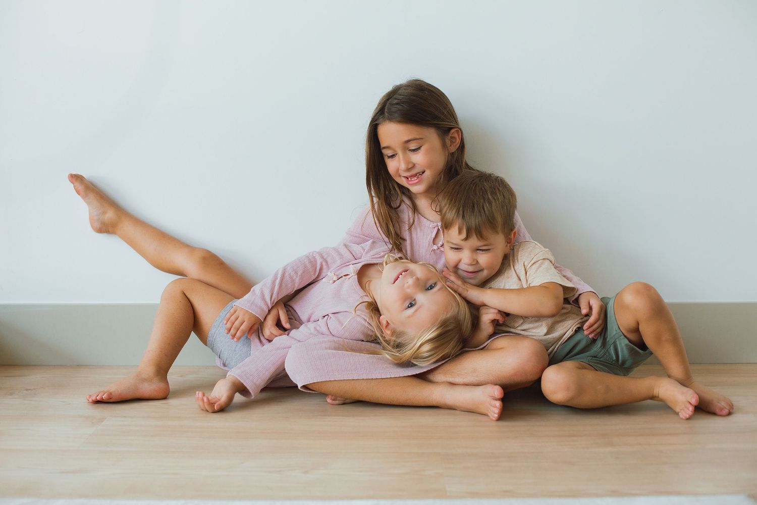 Three happy barefoot children sitting together on a wooden floor against a light gray wall.