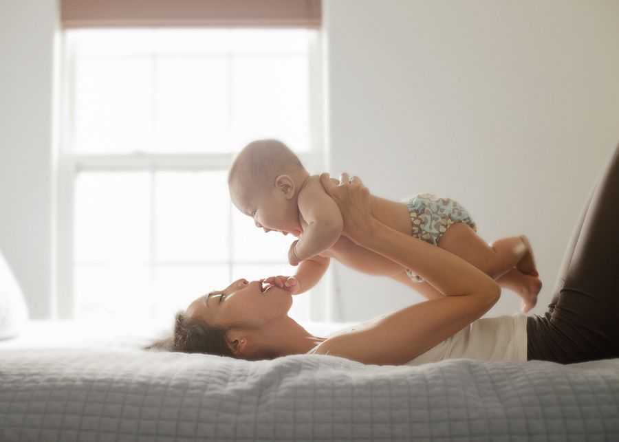 Parent lifting baby playfully while lying on bed in sunlit room.