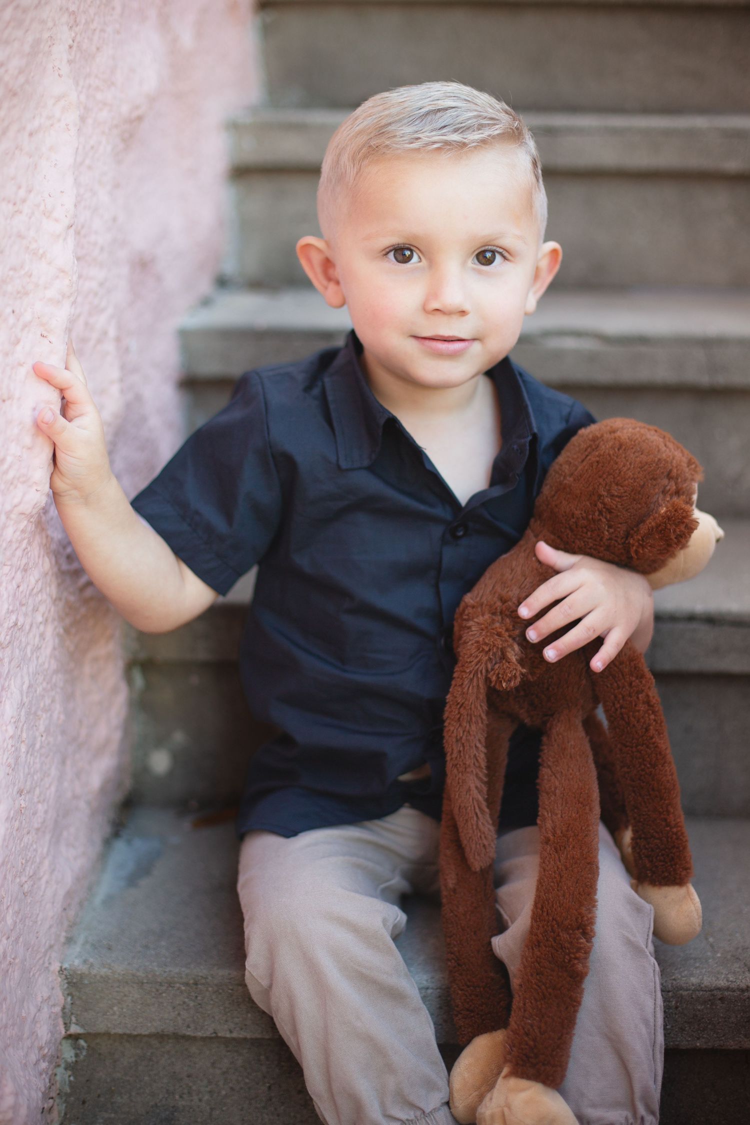 Young child in navy shirt sits on steps holding brown stuffed monkey toy while leaning against pink wall.
