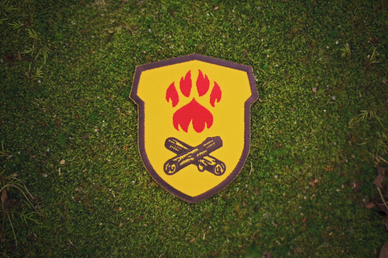 Yellow shield emblem with red paw print and crossed sticks against grass background.