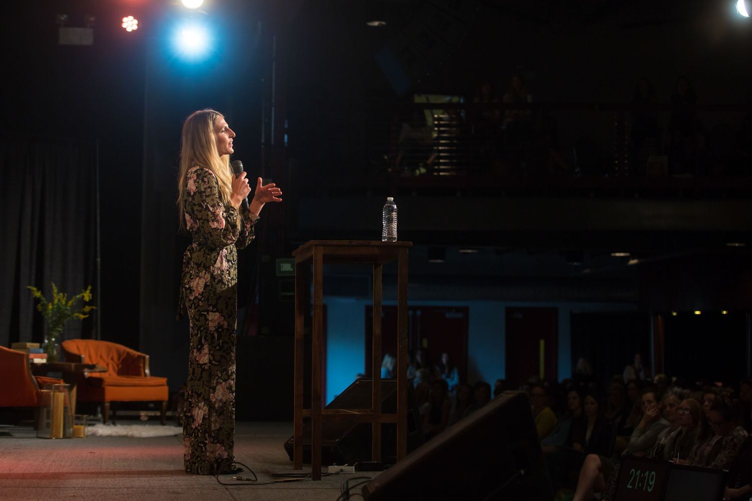 Performer speaking on stage with dramatic lighting and audience in background.