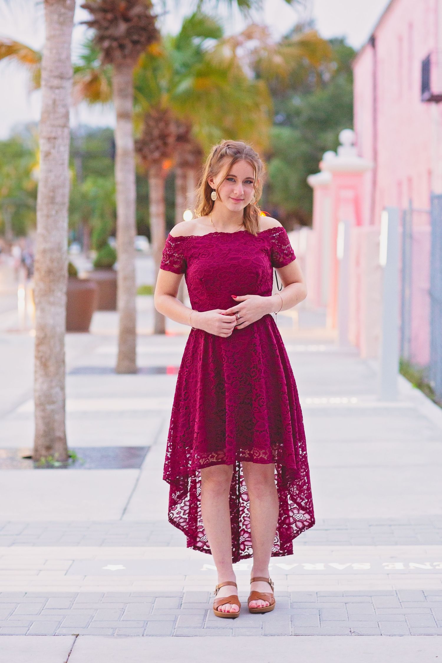 Person in burgundy lace dress standing on sidewalk with palm trees and pink building.