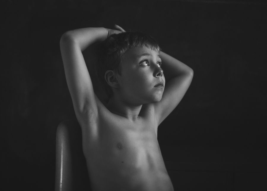Dramatic black and white portrait of shirtless child with hands behind head looking upward.