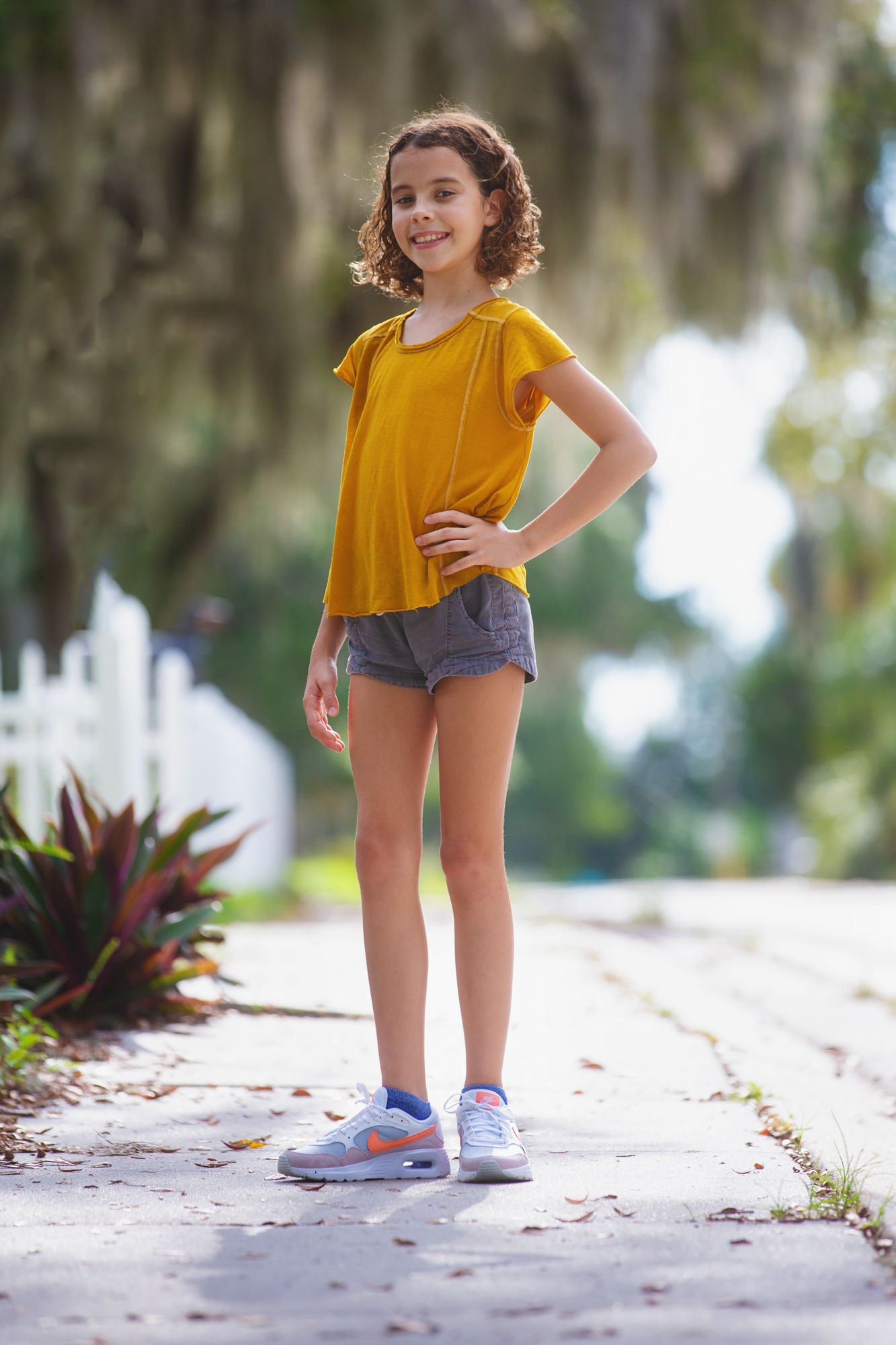 A young person in a yellow top and shorts stands confidently on a sunny sidewalk.