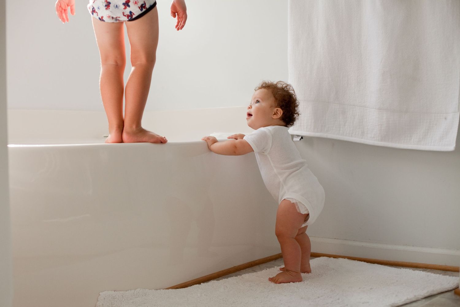 Baby in white onesie learning to stand against wall while adult watches nearby.