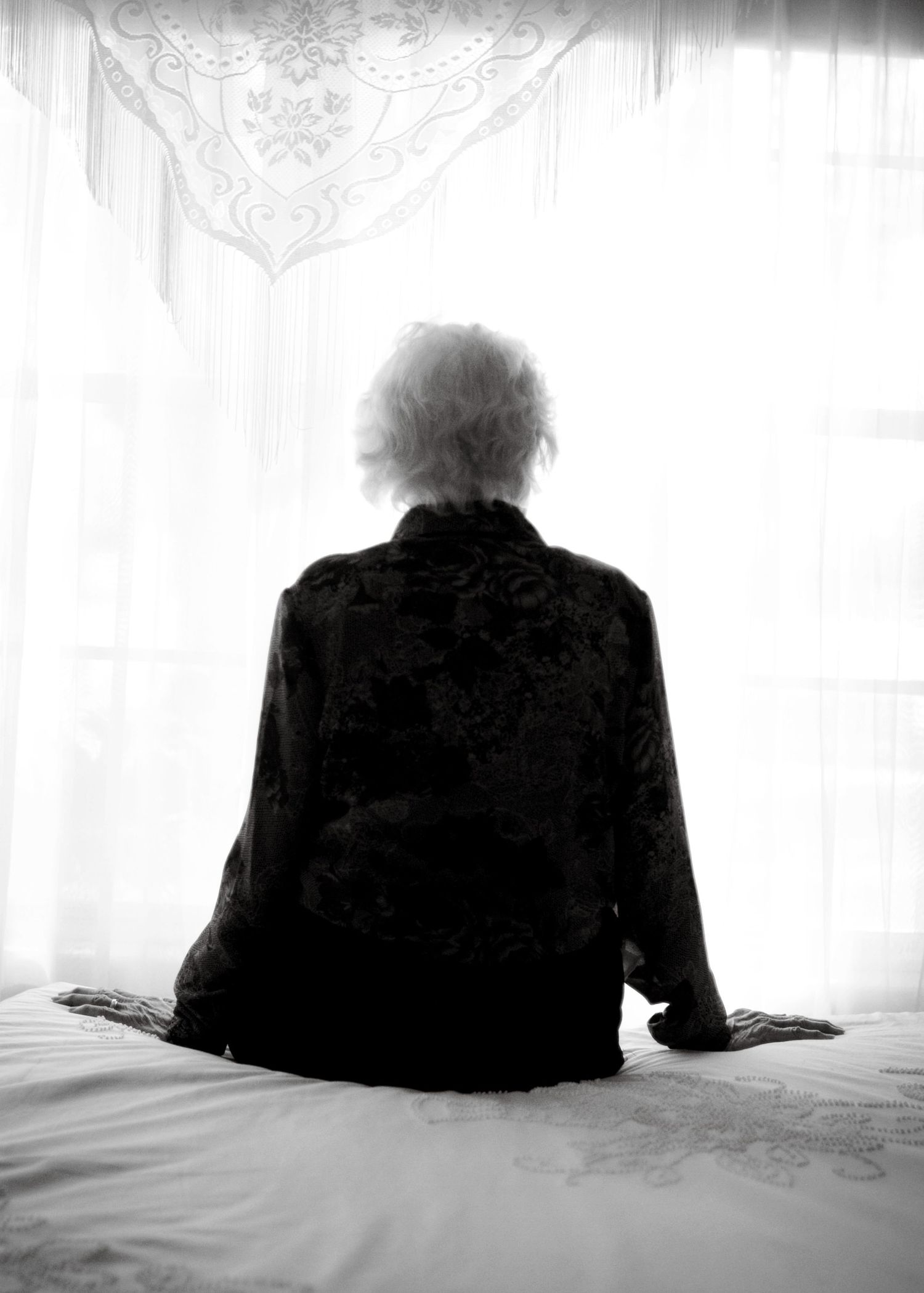 A silhouetted figure sits alone on a bed facing a curtained window in moody lighting.