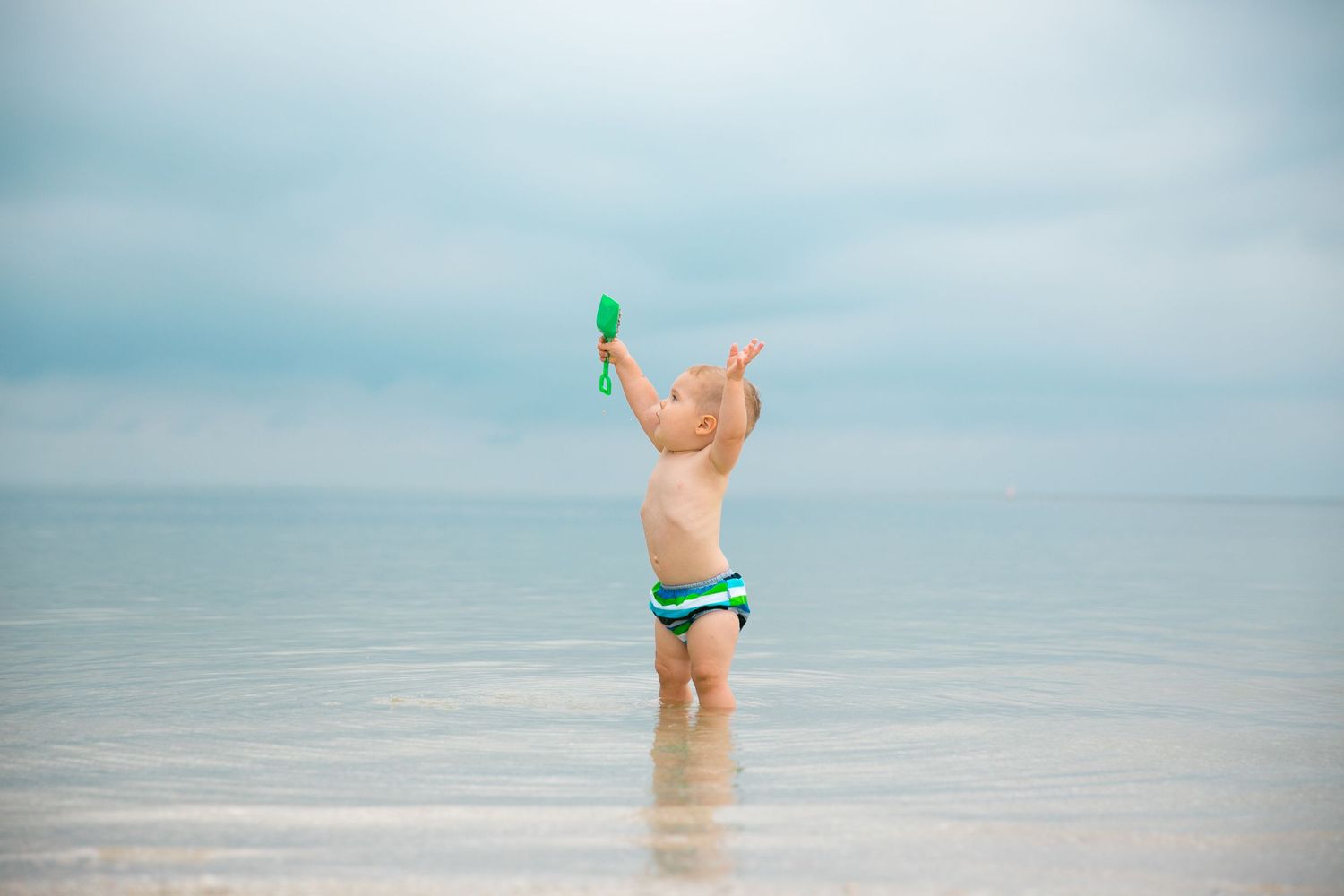 Small figure playing with green toy in shallow ocean water under cloudy sky.