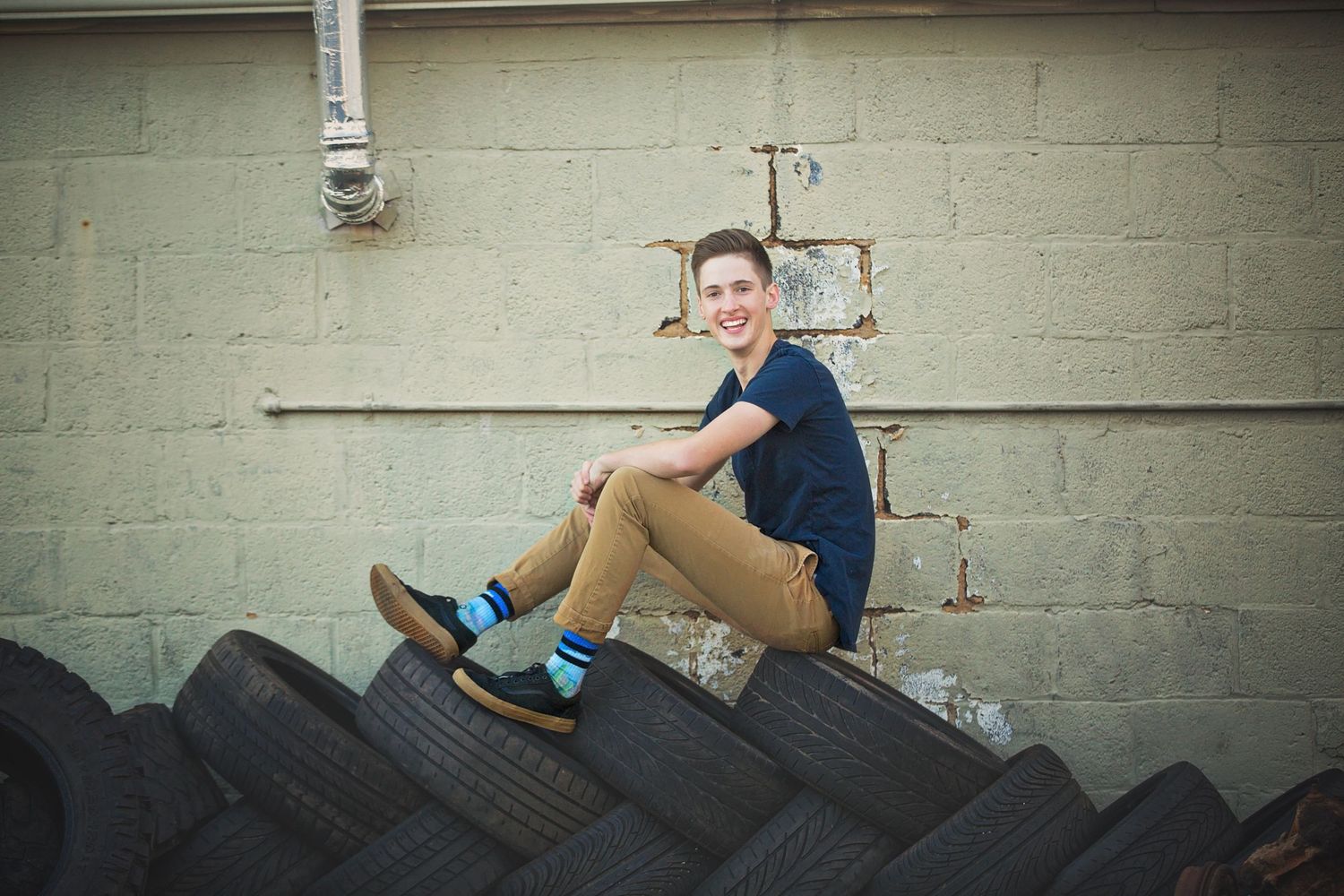 A person sits casually on a pile of tires against an industrial wall background.