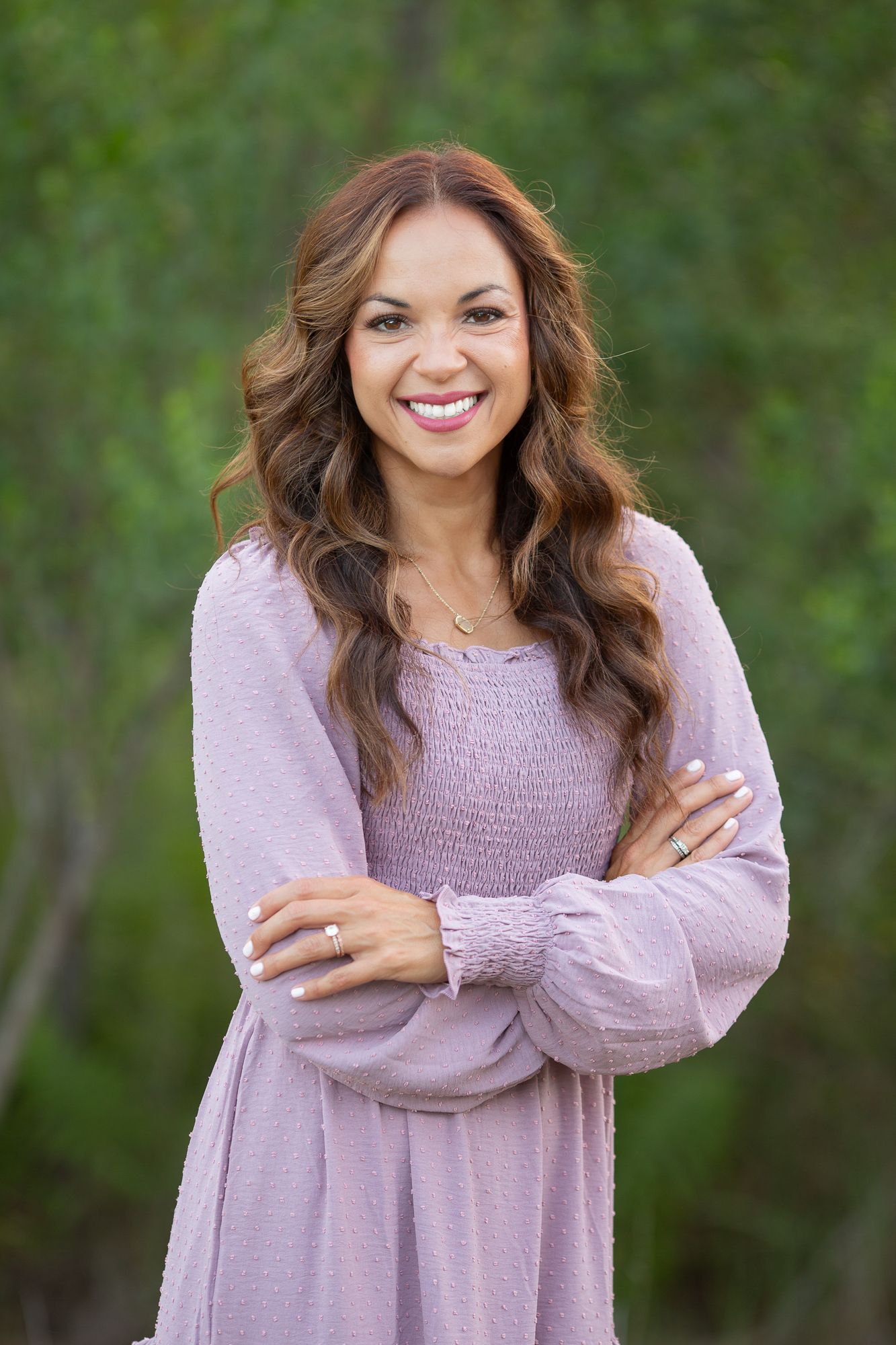 Smiling woman with wavy brown hair in lavender dress, arms crossed, posing outdoors with green foliage background.