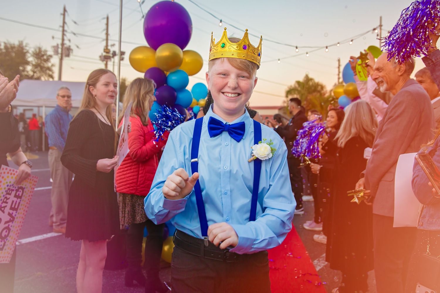 Smiling teen in blue shirt, bow tie, suspenders, and gold crown celebrates at an outdoor prom or homecoming event.