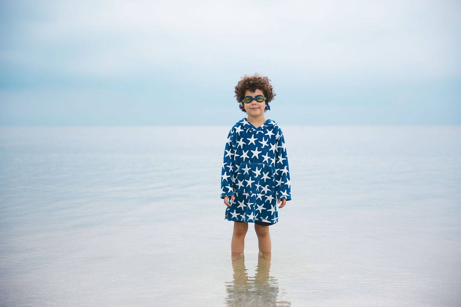 Child in a blue floral dress standing in shallow ocean water on a cloudy day.