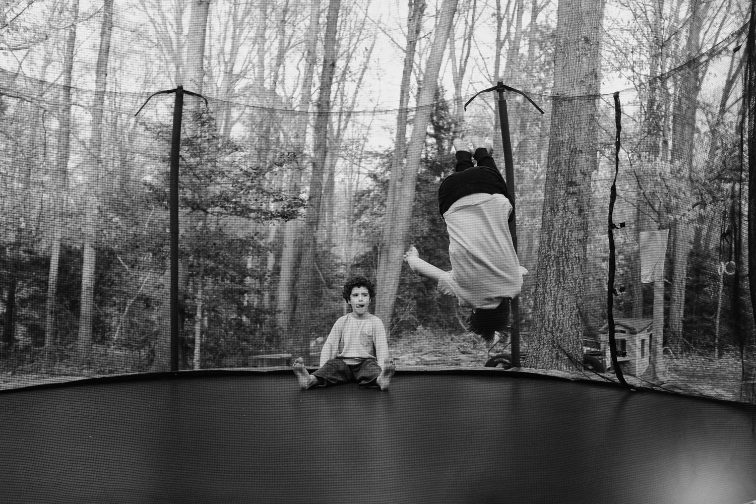 Two people jumping on a trampoline in a wooded backyard setting captured in black and white.