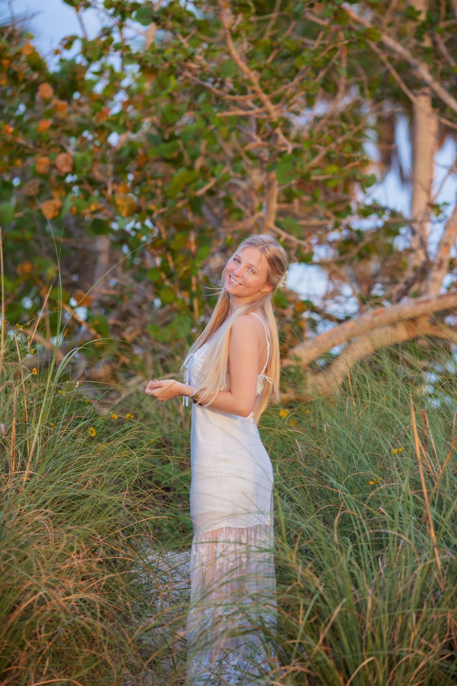 Smiling blonde woman in white maxi dress standing among tall grass and tropical trees outdoors.