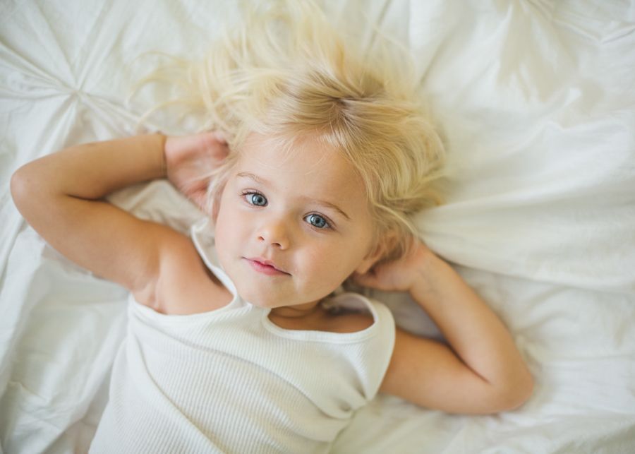 Young child with blonde hair lying on white bedding looking at camera.