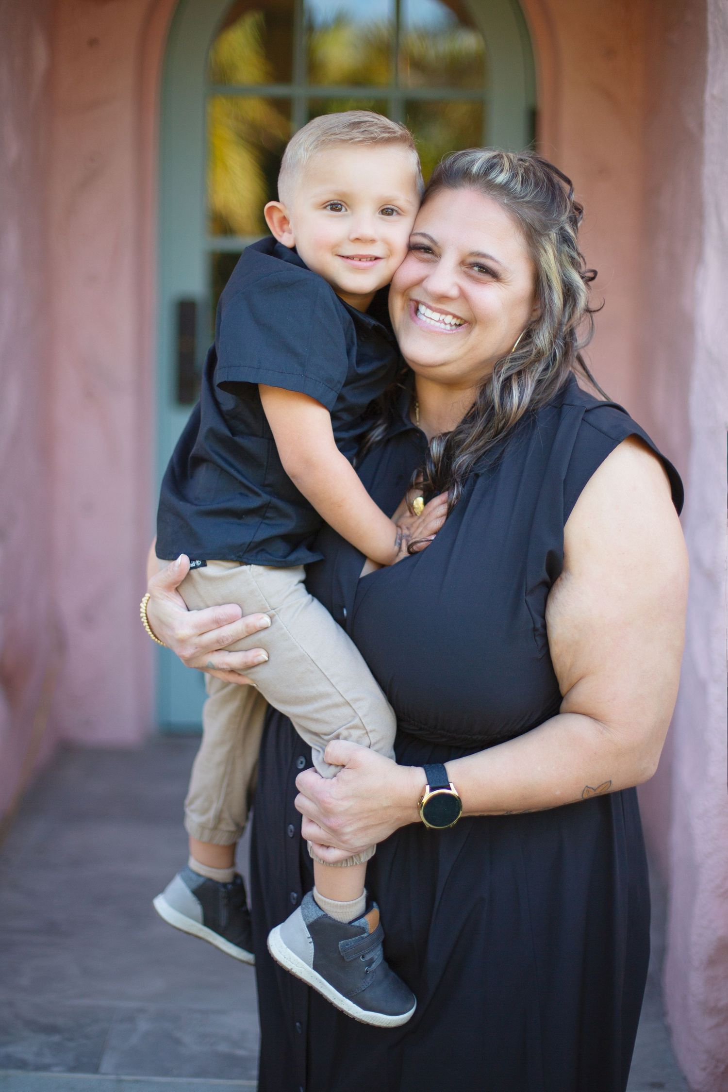 A woman in a black dress holds a young child in front of a pink-hued wall with an arched doorway.