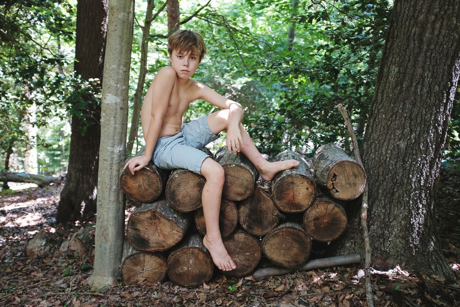 A barefoot child sits casually on a stack of cut logs in a wooded area.