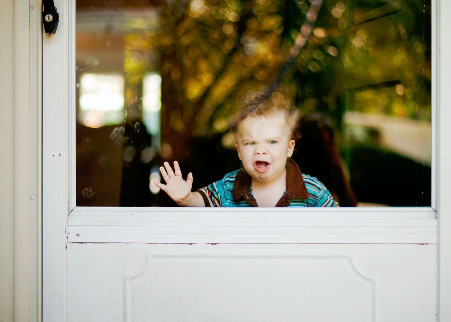 Child making silly face and waving through glass door window.