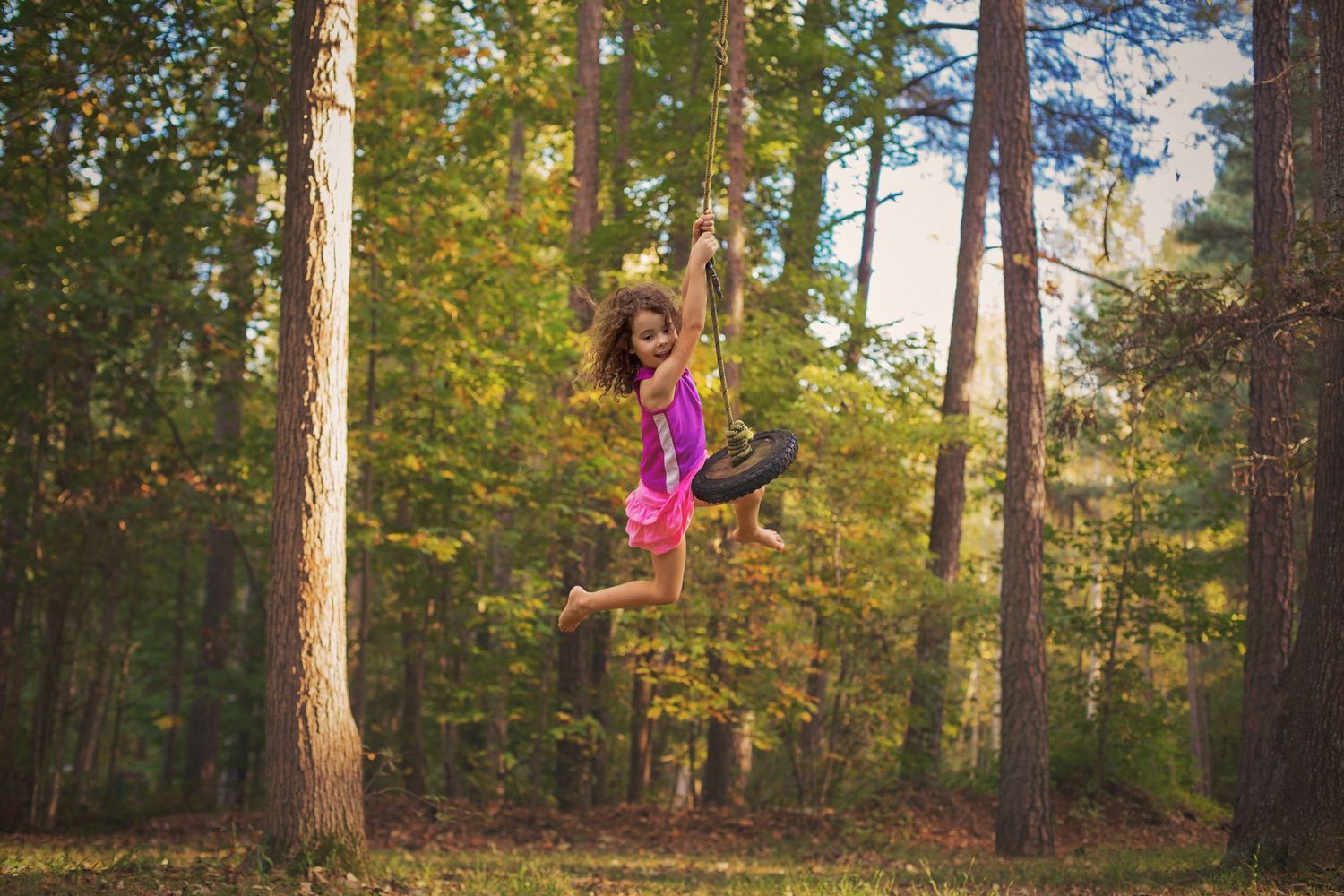Dancer in pink dress leaping gracefully through a sunlit forest setting.