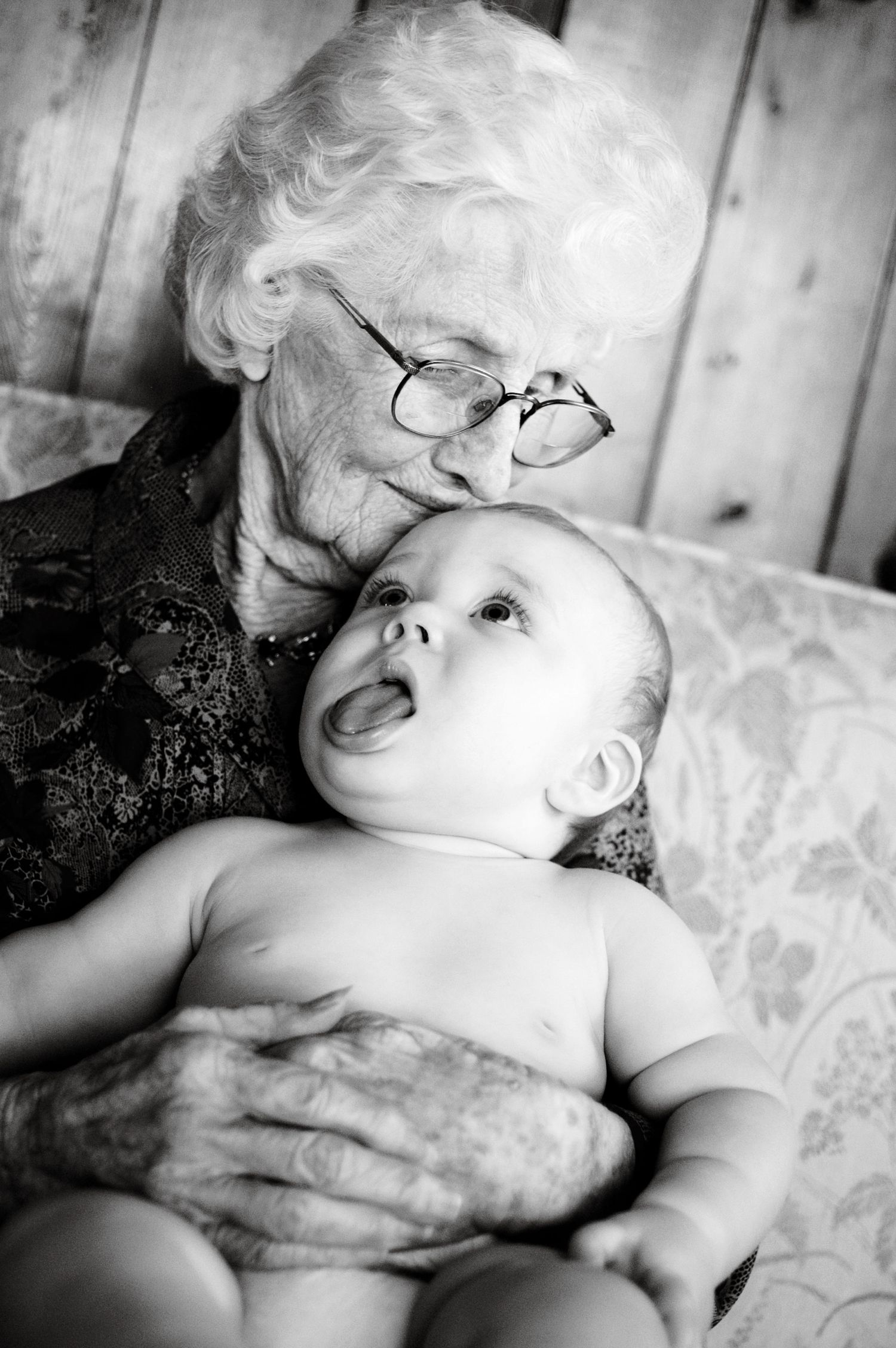 An elderly person wearing glasses holds a smiling baby in a tender black and white portrait.