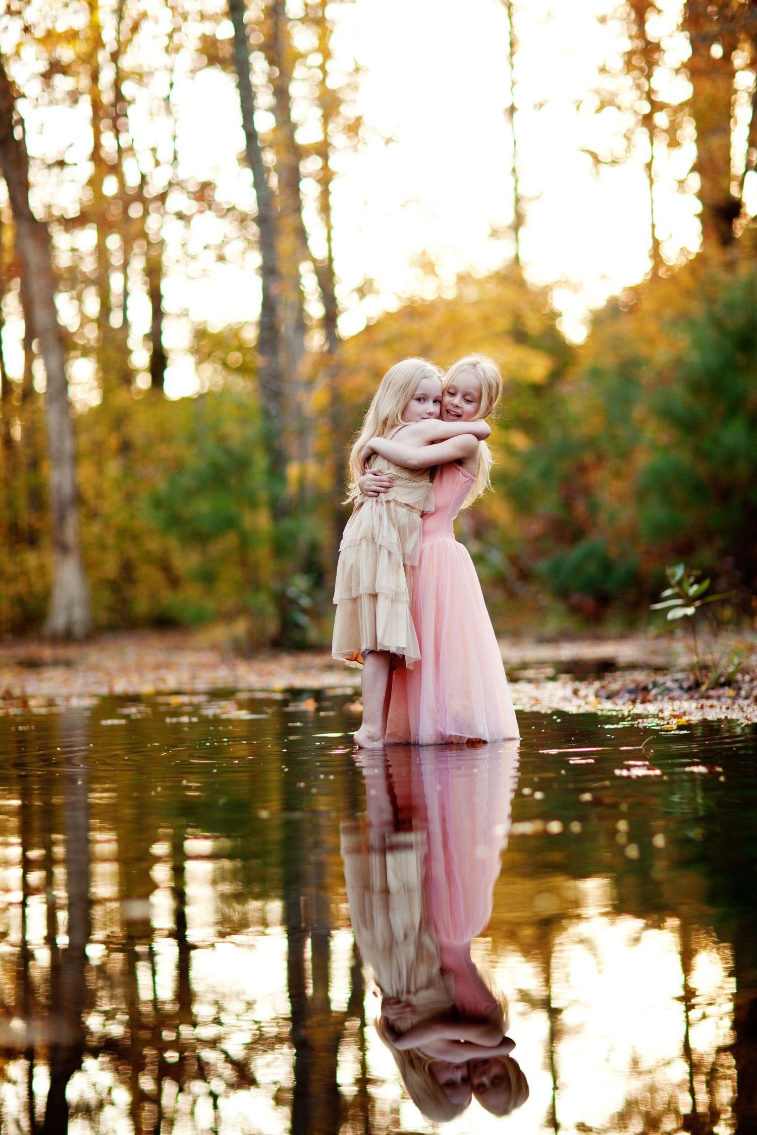 Two people embracing in flowing dresses reflected in still water during golden sunset.